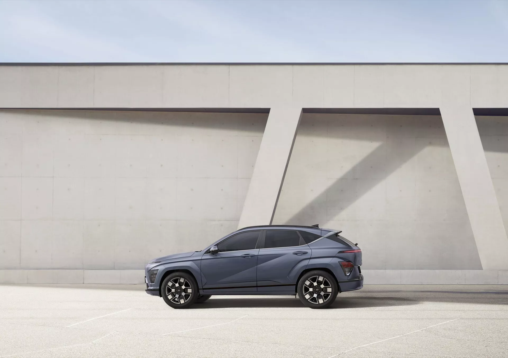 Hyundai Kona drugiej generacji 2023 w widoku z profilu bocznego prezentujący premium felgi aluminiowe, konstrukcję z czterema drzwiami, kierunkowskazy zintegrowane z lusterkami bocznymi, i eleganckie przyciemniane szyby, umieszczone na tle modernistycznym pod jasnym światłem dziennym