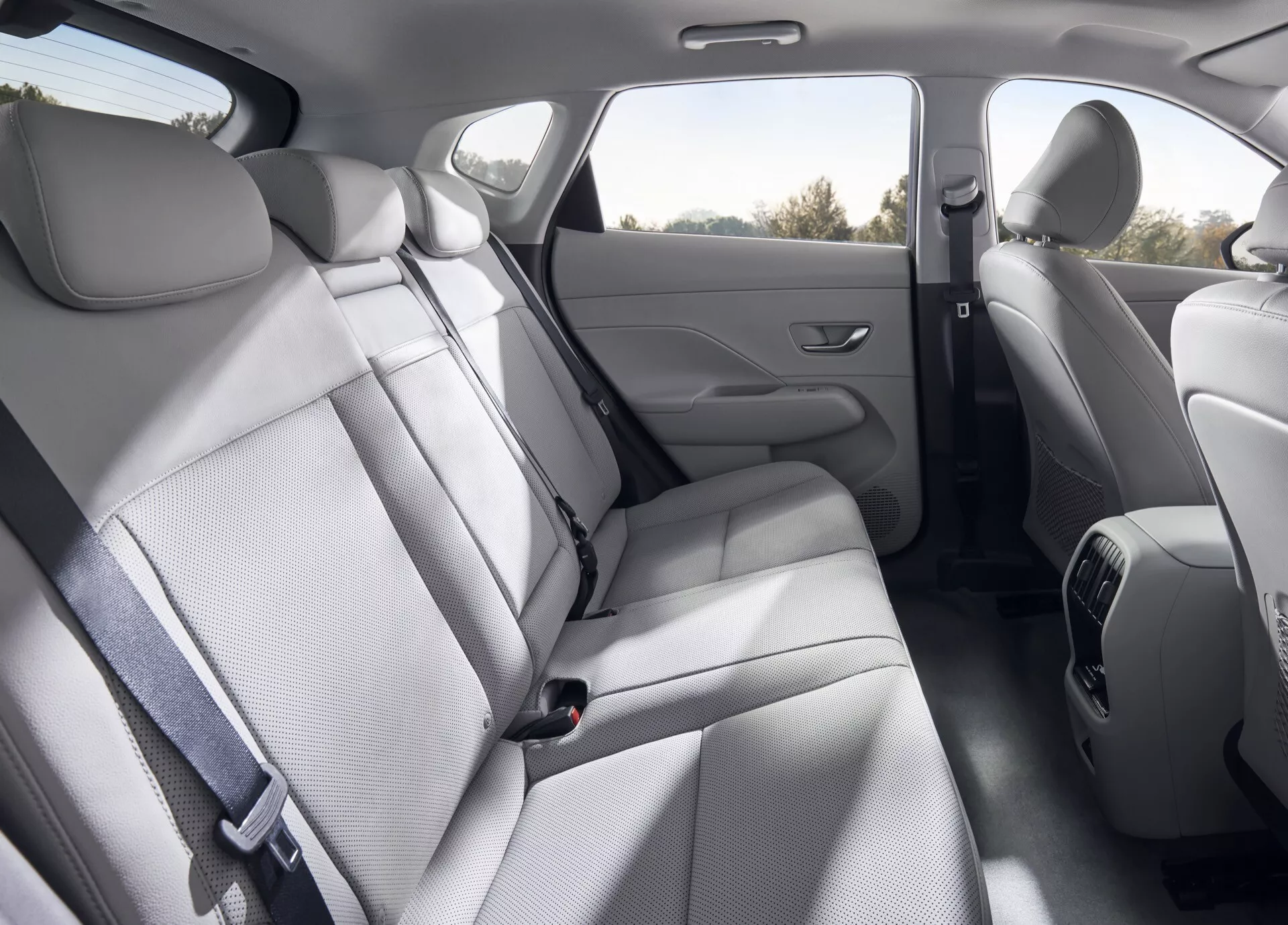 Wnętrze tylnej części pasażerskiej Hyundai Kona 2 generacji 2023 z siedzeniami ławkowymi w jasnoszarej tkaninie, obszerną przestrzenią na nogi, zintegrowanymi podłokietnikami w drzwiach i tylnymi nawiewami.