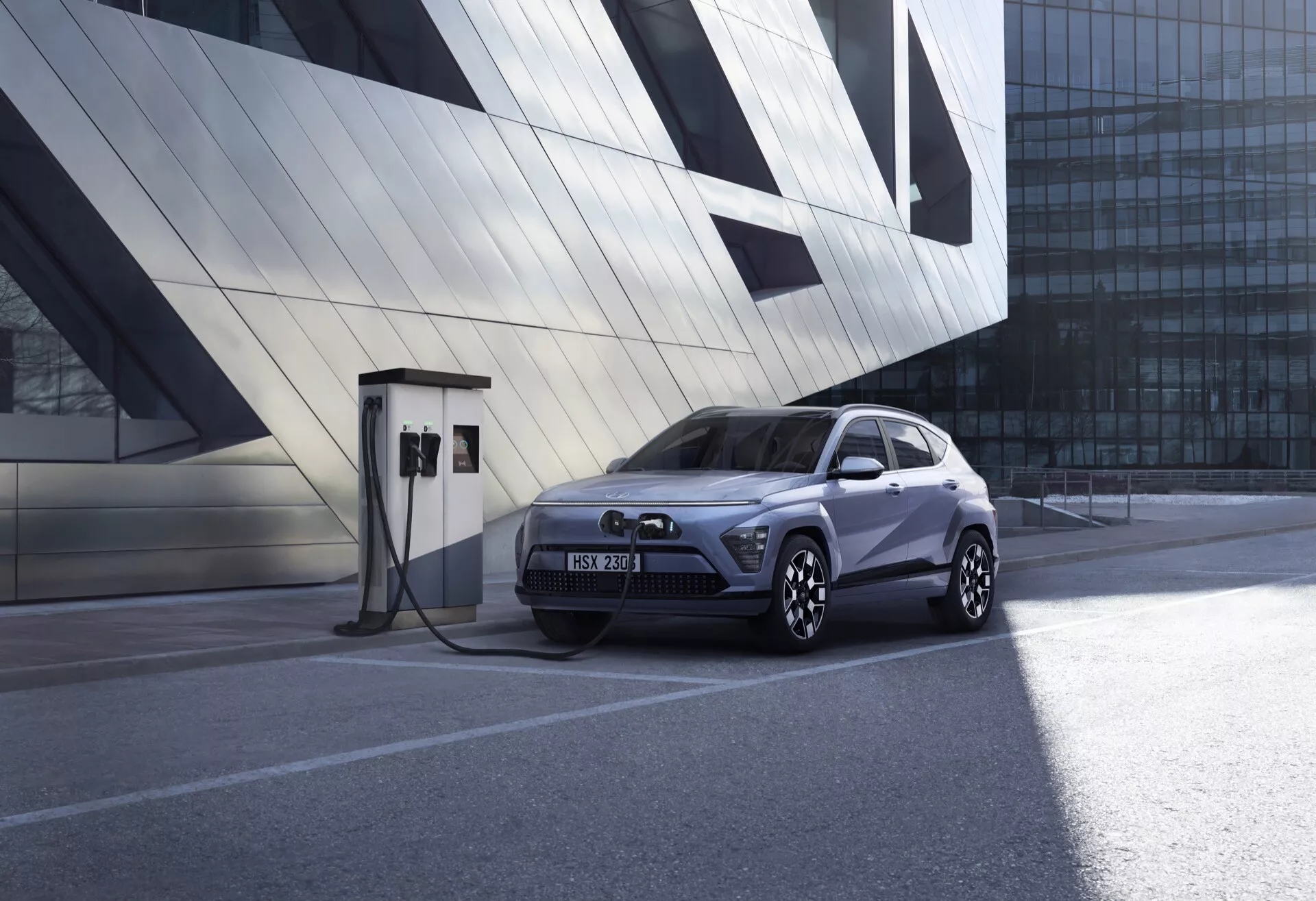 Hyundai Kona 2023 zaparkowany przy stacji ładowania pojazdów elektrycznych, prezentujący niski przedni trzy czwarte kąt podkreślający nowoczesny design SUV, charakterystyczne światła LED, premiumowe felgi aluminiowe o wielu szprychach i podkreśloną technologię pojazdów elektrycznych w miejskim otoczeniu