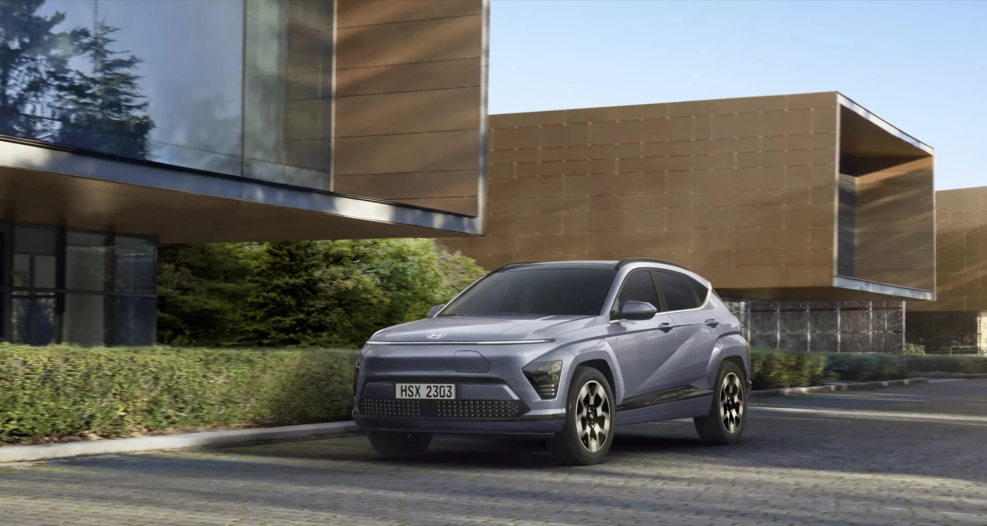 Hyundai Kona 2. generacji 2023 kompaktowy SUV w miękkim szarym kolorze, pokazany na asfalcie przed budynkiem o nowoczesnej architekturze, z wysokim prześwitem, zamkniętą atrapą chłodnicy dla modeli elektrycznych lub hybrydowych, eleganckimi reflektorami LED i felgami ze stopu