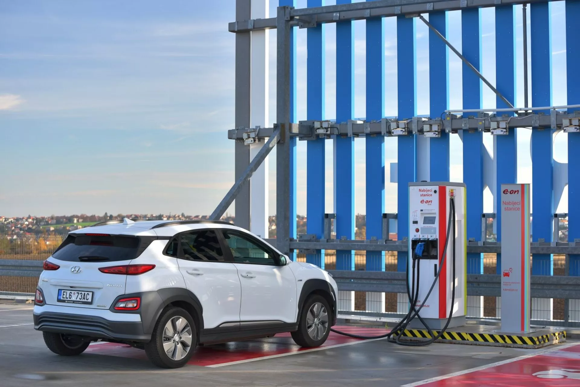 Biały elektryczny SUV Hyundai Kona 2021, ukazany z tyłu z perspektywy trzech kwart na zewnątrznej stacji ładowania z tłem czystego nieba