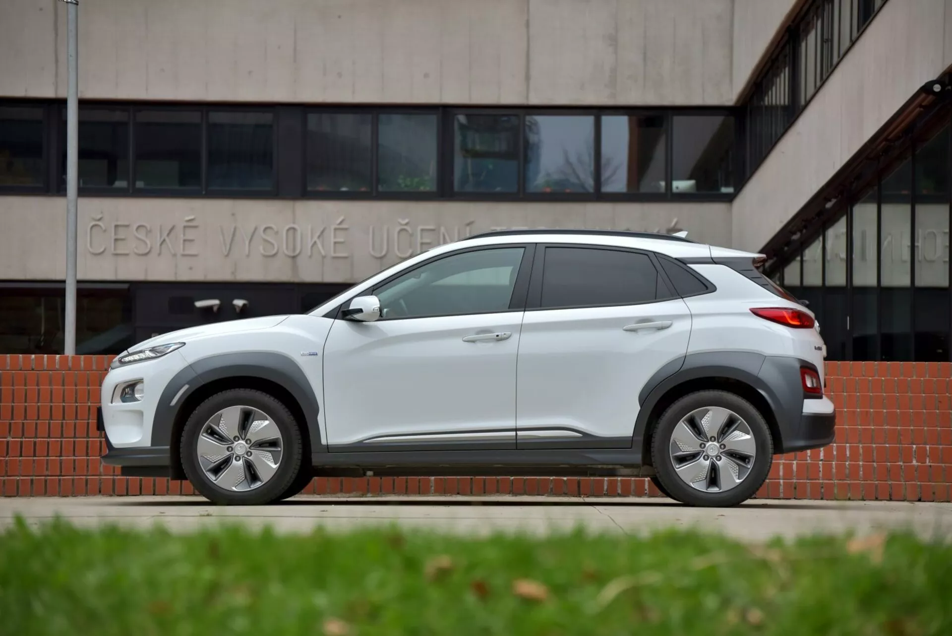 Profil boczny Hyundai Kona Elektro 2021 w jasnoszarym kolorze, podkreślający elegancki design z nadkolami i progami w kolorze nadwozia oraz nowoczesne cechy jak koła ze stopów lekkich i antena w kształcie płetwy rekina, przed budynkiem z napisem 'ČESKÉ VYSOKÉ UČENÍ', co podkreśla jego przydatność do jazdy miejskiej i terenowej