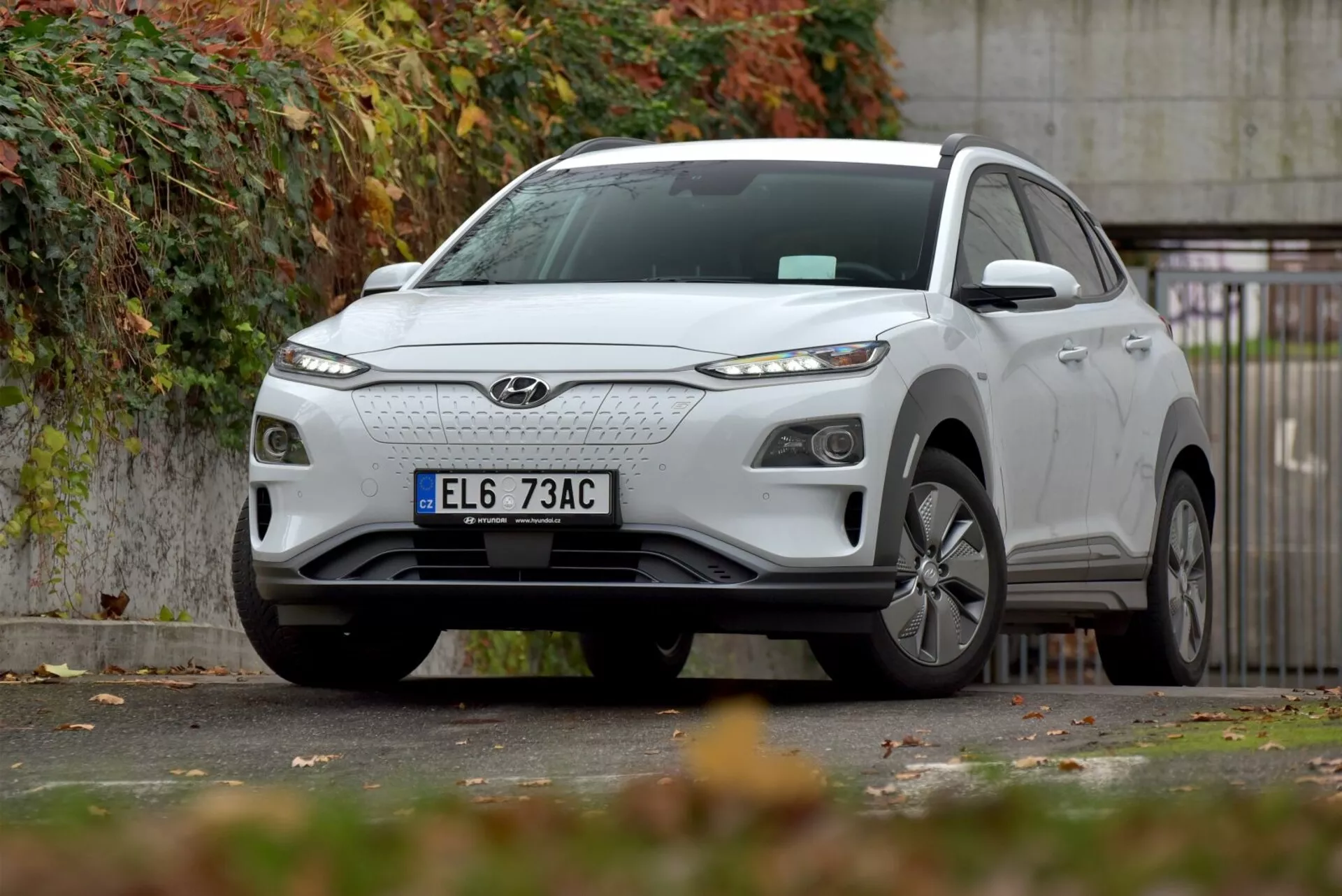 Elektryczny kompaktowy SUV Hyundai Kona 2021 zaparkowany przy miejskiej drodze, prezentujący przedni grill, eleganckie reflektory LED, aluminiowe koła i rzeźbiony design nadwozia z naciskiem na lewą stronę przednią, na tle jesiennej scenerii