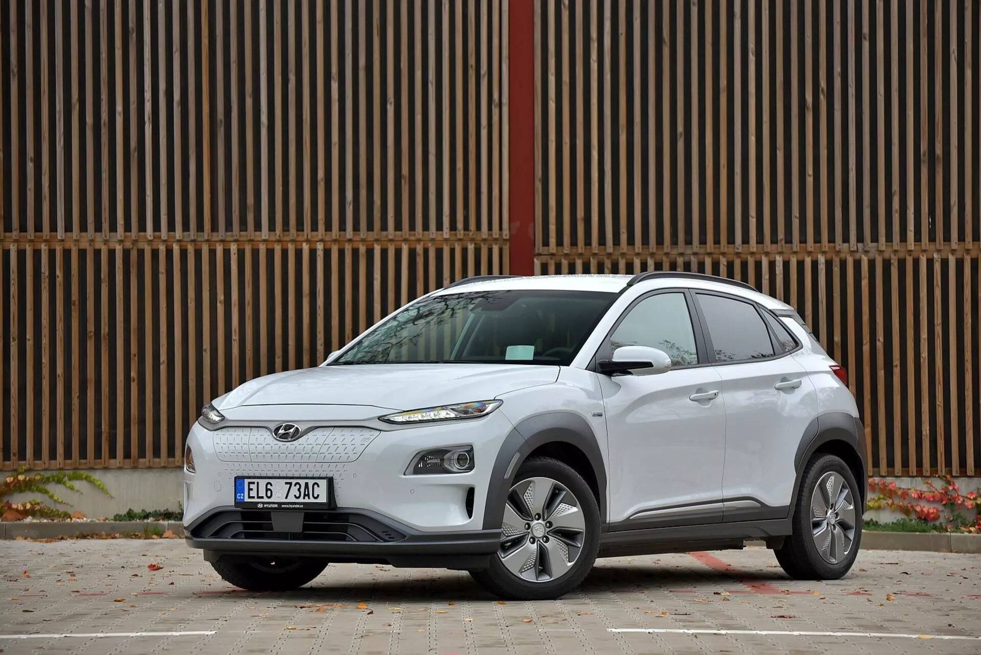 Elektryczny kompaktowy SUV Hyundai Kona 2021 w kolorze białym, prezentujący swój nowoczesny design z profilu bocznego ujęcia trzech czwartych, z większością zamkniętym grillem, ostrymi reflektorami, felgami aluminiowymi, zintegrowanymi kierunkowskazami w lusterkach bocznych w kolorze nadwozia, standardowymi klamkami drzwi, przyciemnianymi szybami dla tylnych okien, sugestią tylnego spoilera, zaparkowany na utwardzonej powierzchni w miejskim otoczeniu.