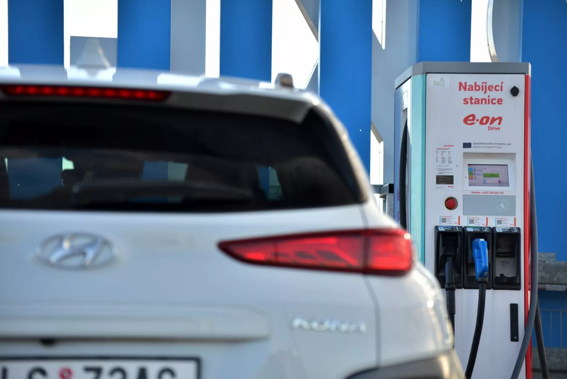 Zbliżenie na tylną prawą część białego elektrycznego pojazdu Hyundai Kona 2021, skupiające się na eleganckim projekcie tylnej lampy, z częściowo widocznym logo Hyundai i nazwą modelu w pobliżu środka tylnej klapy. Częściowo widoczna tablica rejestracyjna na dole lewej strony tylnej klapy wskazuje na staranne zachowanie prywatności. Na tle znajduje się stacja ładowania pojazdów elektrycznych E.ON, podkreślając elektryczny charakter samochodu na rozmytym tle, co wskazuje na elektryczną mobilność i technologię zrównoważonego transportu.