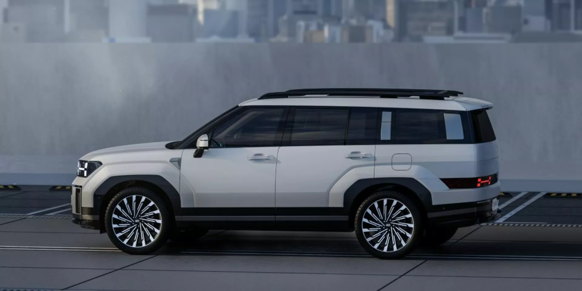 Profil boczny SUV-a Hyundai Santa Fe 2024, ukazujący jego pudełkowaty, ale nowoczesny design z wyraźnymi liniami i krawędziami. Widoczne cechy obejmują boczne drzwi, okna, lusterka boczne, duże wieloramienne koła aluminiowe, dwukolorowy lakier z jasną karoserią i ciemniejszym dachem oraz dolnymi listwami, wysokie prześwity, podesty i relingi dachowe, wszystko na tle niewyraźnego, rozmytego miejskiego otoczenia.
