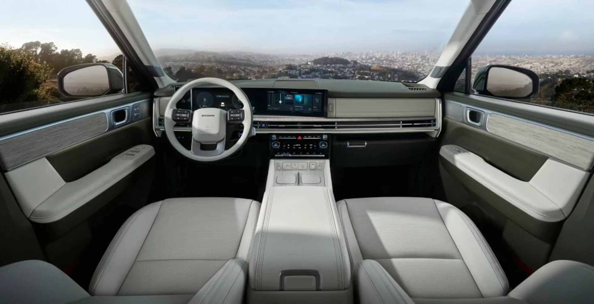 Wnętrze Hyundai Santa Fe 2024 (5. generacja), ukazujące przednią część kabiny z nowoczesną, minimalistyczną deską rozdzielczą, skórzaną dwuramienną kierownicą i dotykowym ekranem systemu multimedialnego. Konsola centralna wyposażona jest w elegancki elektroniczny selektor biegów, uchwyty na napoje i schowki. Przednie fotele są tapicerowane jasną skórą, zapewniając komfort i ergonomię. Wnętrza drzwi zawierają podłokietniki, panele kontrolne do elektrycznych szyb i dodatkowe schowki, wszystkie wykonane z wysokiej jakości materiałów. Cały design emanuje luksusem, technologią i przestrzennością.