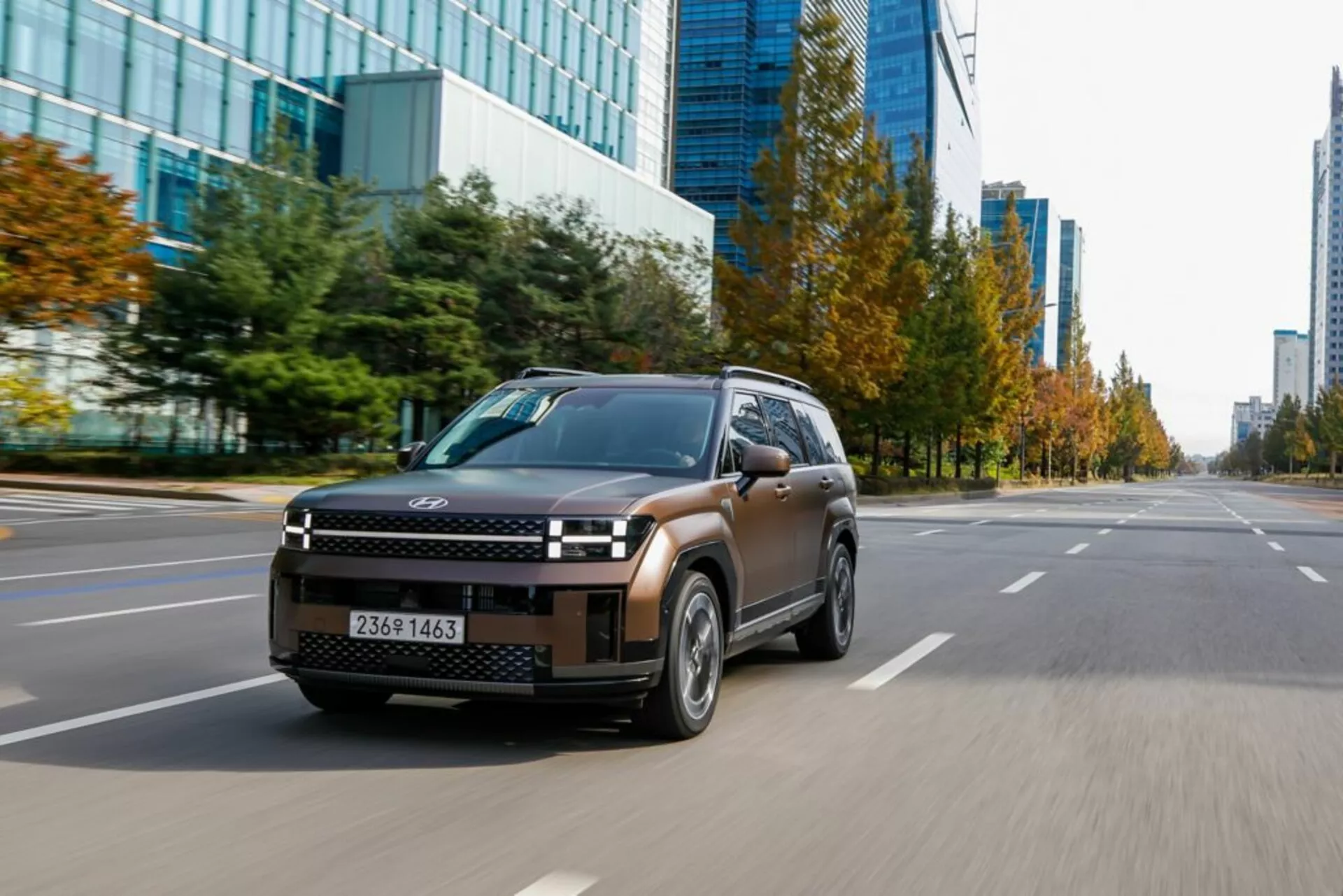 Obraz modelu Hyundai Santa Fe 2024 piątej generacji widziany z trzech czwartych z przodu z lewej strony, uchwycony pod niskim kątem, pokazujący elegancką przednią kratę SUV-a, reflektory LED, maskę, przednie błotniki, przednią szybę, drzwi boczne i boczne lusterka. Samochód porusza się po miejskiej drodze z wieloma pasami ruchu i komercyjnymi budynkami w tle. Aerodynamiczne felgi i częściowo rozmyta tablica rejestracyjna podkreślają ruch i osiągi pojazdu w miejskim środowisku.