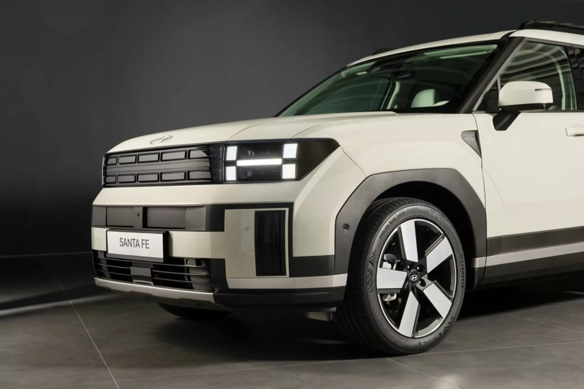 Zbliżenie z niskiego kąta na Hyundai Santa Fe 2024 (5. generacja) z naciskiem na przedni lewy kwartalny, podkreślające nowoczesny design przedniego grilla, segmentowe reflektory ze specyficznymi światłami do jazdy dziennej, wyrzeźbioną maskę, wieloramienne aluminiowe koło, widoczny bieżnik opony, grilla z logo, linię charakterystyczną na błotniku, aerodynamiczne przednie lewe drzwi i lusterko boczne, oraz neutralny kolor pod oświetleniem studyjnym.