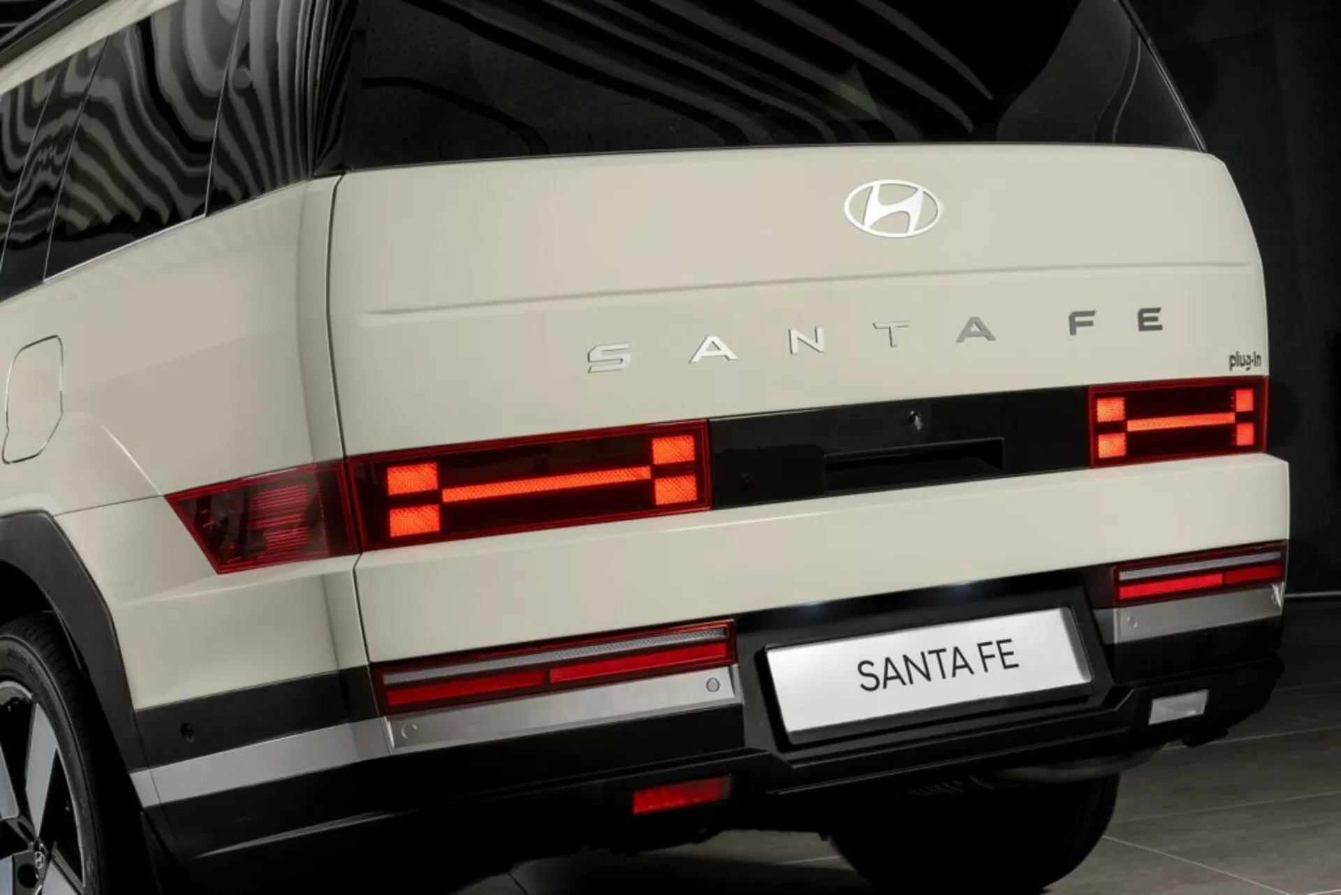 Zbliżenie tylnego widoku Hyundai Santa Fe 2024 (5. generacja), zrobione z poziomu ziemi. Na zdjęciu widać podświetlone poziome, segmentowe światła tylne, srebrną listwę, emblemat Hyundai i markę 'SANTA FE'. Widoczne są tylna wycieraczka, obszar tablicy rejestracyjnej i potencjalne światła przeciwmgielne lub odblaski na poziomie zderzaka. Przedstawione w środowisku wewnętrznym, sugerując dobrze utrzymane, nowoczesne pojazdy, prawdopodobnie używane do celów promocyjnych.