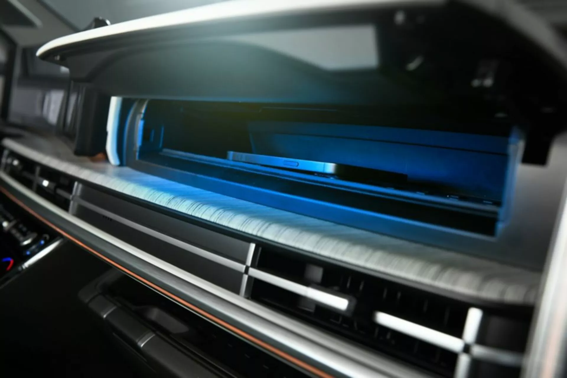 Zbliżenie szczegółów systemu wentylacji wewnątrz deski rozdzielczej Hyundai Santa Fe 2024 5. generacji, oglądane pod nieco wyższym kątem. Poziome listwy wentylacyjne prawie zamknięte z małą głębią ostrości, tworzącą rozmycie na pierwszym planie i tle. Nowoczesne wykończenie z metalu szczotkowanego i potencjalnie dotykowe elementy sterujące, wzbogacone niebieskim oświetleniem ambientowym, nadające wnętrzu high-tech i premium. Kontrastujący materiał wokół wentylacji sugeruje luksusowe wykończenie, prawdopodobnie drewno lub fornir drewnopodobny. Brak widocznych pokręteł/przycisków wskazuje na nowoczesny dotykowy system HVAC. Zdjęcie wnętrza skupia się na komforcie, wygodzie i zaawansowanych funkcjach sterowania klimatem.