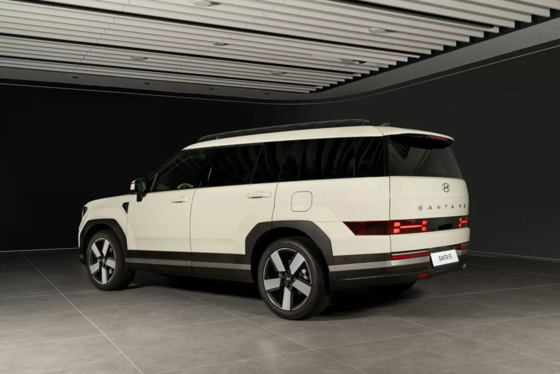 Widok tylno-boczny SUV Hyundai Santa Fe 2024 (5. generacja) pokazany w neutralnym otoczeniu wewnętrznym. Widoczne są tylne światła z poziomymi liniami, tylna szyba, zderzak i miejsce na tablicę rejestracyjną. Elegancki profil boczny ukazuje klamki drzwi, linie konturu i częściowo widoczny panoramiczny dach. Nowoczesne funkcje obejmują duże felgi aluminiowe wieloramienne, lusterko boczne z wbudowanymi kierunkowskazami i przyciemniane szyby. Jasny kolor SUV-a i kontrolowane otoczenie sugerują, że jest to luksusowy model z zaawansowaną technologią.