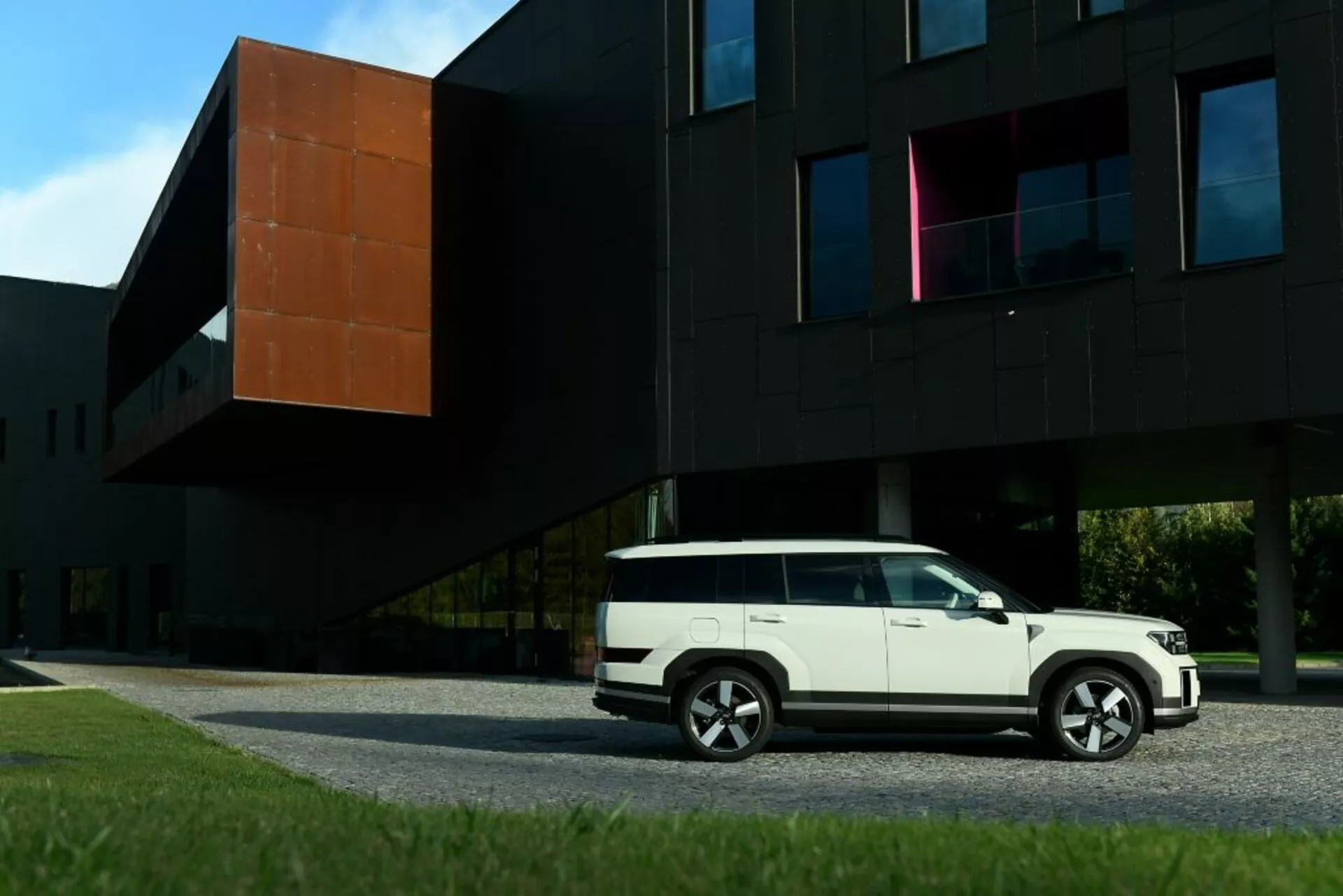 Widok z boku Hyundai Santa Fe 2024 (5. generacja) w białym kolorze, zaparkowanego przed nowoczesnym czarnym budynkiem z pomarańczową częścią. Obraz przedstawia linie nadwozia SUV-a, boczne drzwi, okna, lusterka boczne i wieloramienne felgi aluminiowe. Kompozycja podkreśla zestawienie nowoczesnego wzornictwa pojazdu ze współczesną architekturą.