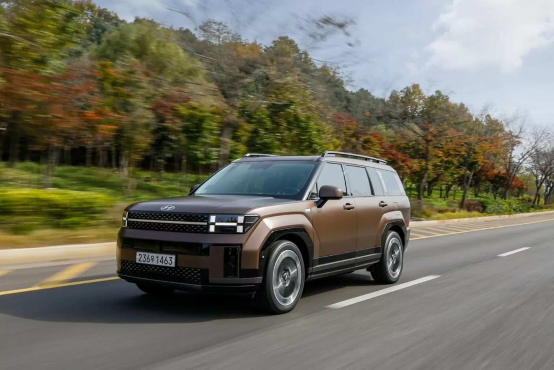 Obraz przedstawia nowoczesne SUV Hyundai Santa Fe 2024 (5. generacja) w ruchu, uchwycone pod kątem trzech czwartych od przodu. Dynamiczne ujęcie podkreśla przedni grill z wyraźnym logo, prostokątne reflektory LED z dziennymi światłami w kształcie litery 'L' oraz masywny przedni zderzak z światłami przeciwmgielnymi. SUV posiada dach panoramiczny, lusterka boczne z wbudowanymi kierunkowskazami, wieloszprychowe felgi aluminiowe i dużą wysokość nadwozia, odpowiednią do warunków miejskich i off-road. Droga w otoczeniu drzew, rozmazanie kół i lekko rozmyte tło sugerują podmiejskie lub wiejskie otoczenie, podkreślając prędkość pojazdu. Stłumiony brązowy kolor i błyszczące wykończenie podkreślają nowoczesny, aerodynamiczny design SUV-a.