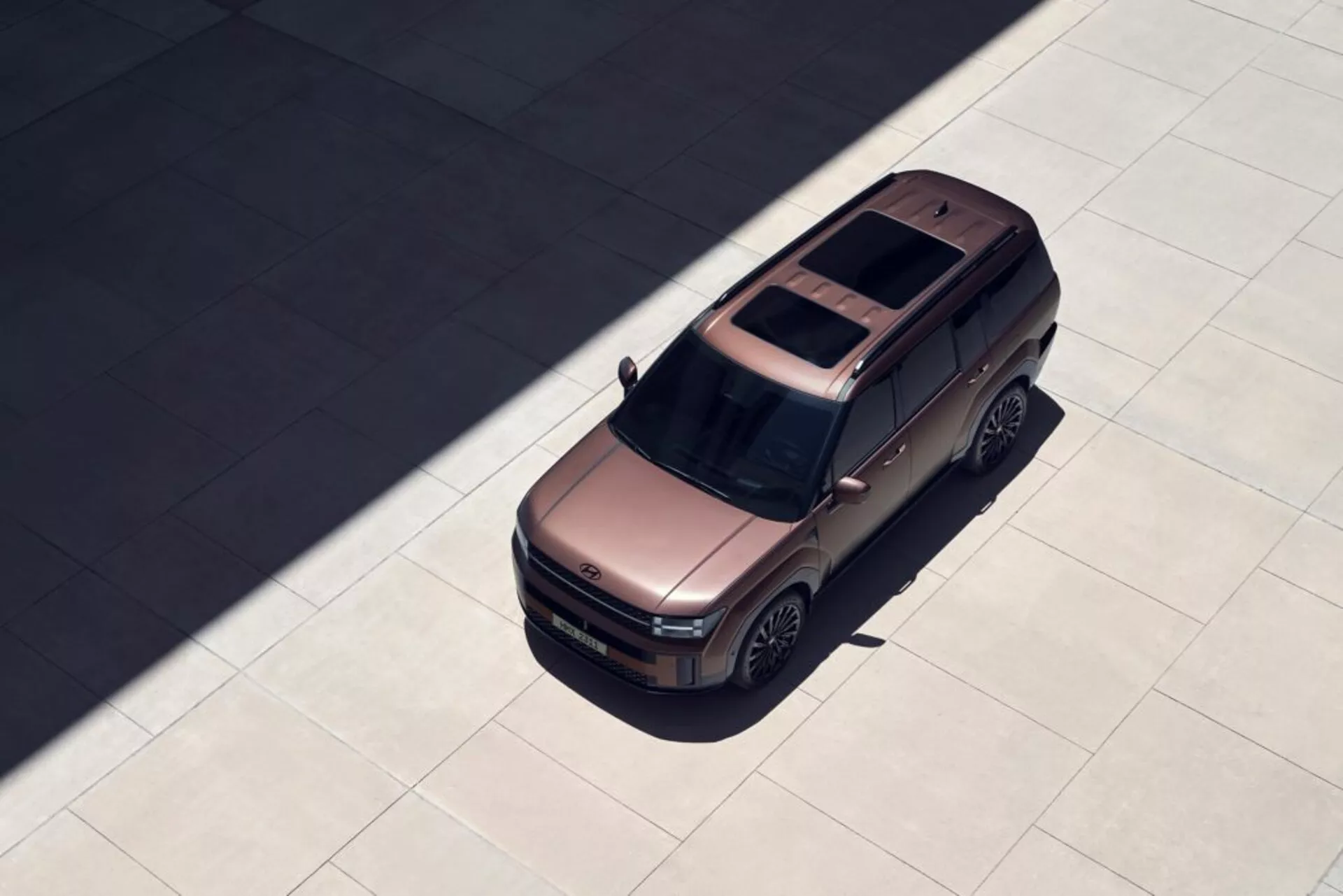 Zdjęcie pod kątem z góry Hyundai Santa Fe 2024 (5. generacji), podkreślające jego masywną, kanciastą konstrukcję, panoramiczny dach, tylną antenę, konturowaną maskę, złożone lusterka boczne, duże aluminiowe koła i cień sugerujący ustawienie oświetlenia o wysokim kontraście. SUV stoi zaparkowany na gładkiej powierzchni, oferując wyraźny widok na jego nowoczesne i wizualnie atrakcyjne cechy.