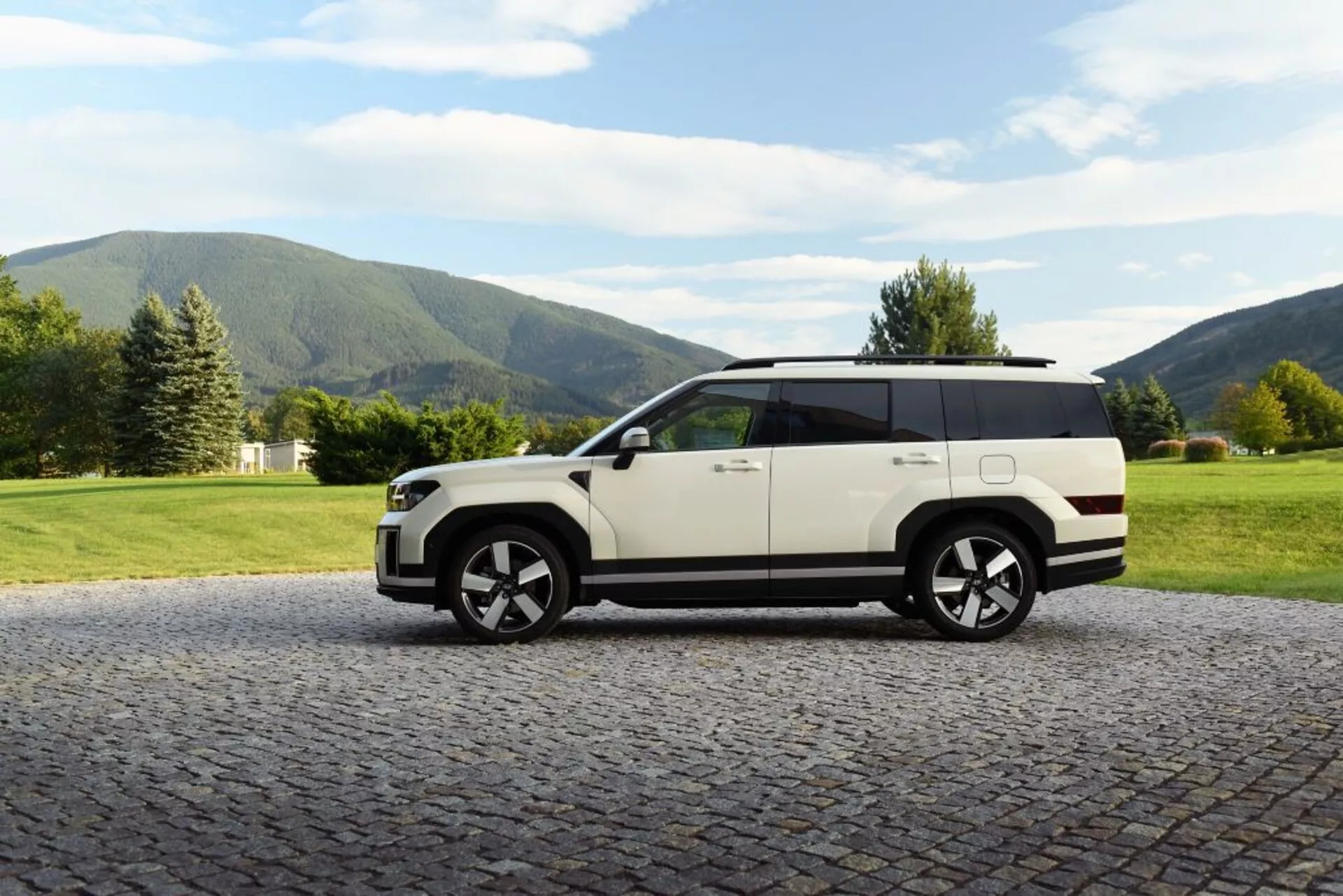 Widok z boku SUV Hyundai Santa Fe 2024 (5. generacja) zaparkowanego na brukowanej nawierzchni z naturalnym krajobrazem w tle. Pojazd ma dwukolorowe malowanie z białym nadwoziem i czarnym dachem, duże felgi aluminiowe, niewidoczne klamki drzwi, co sugeruje technologię chowaną, duże okna, relingi dachowe i wysokie prześwity. Idealny do prezentacji designu samochodowego, luksusu, zaawansowanej technologii i terenowych możliwości.