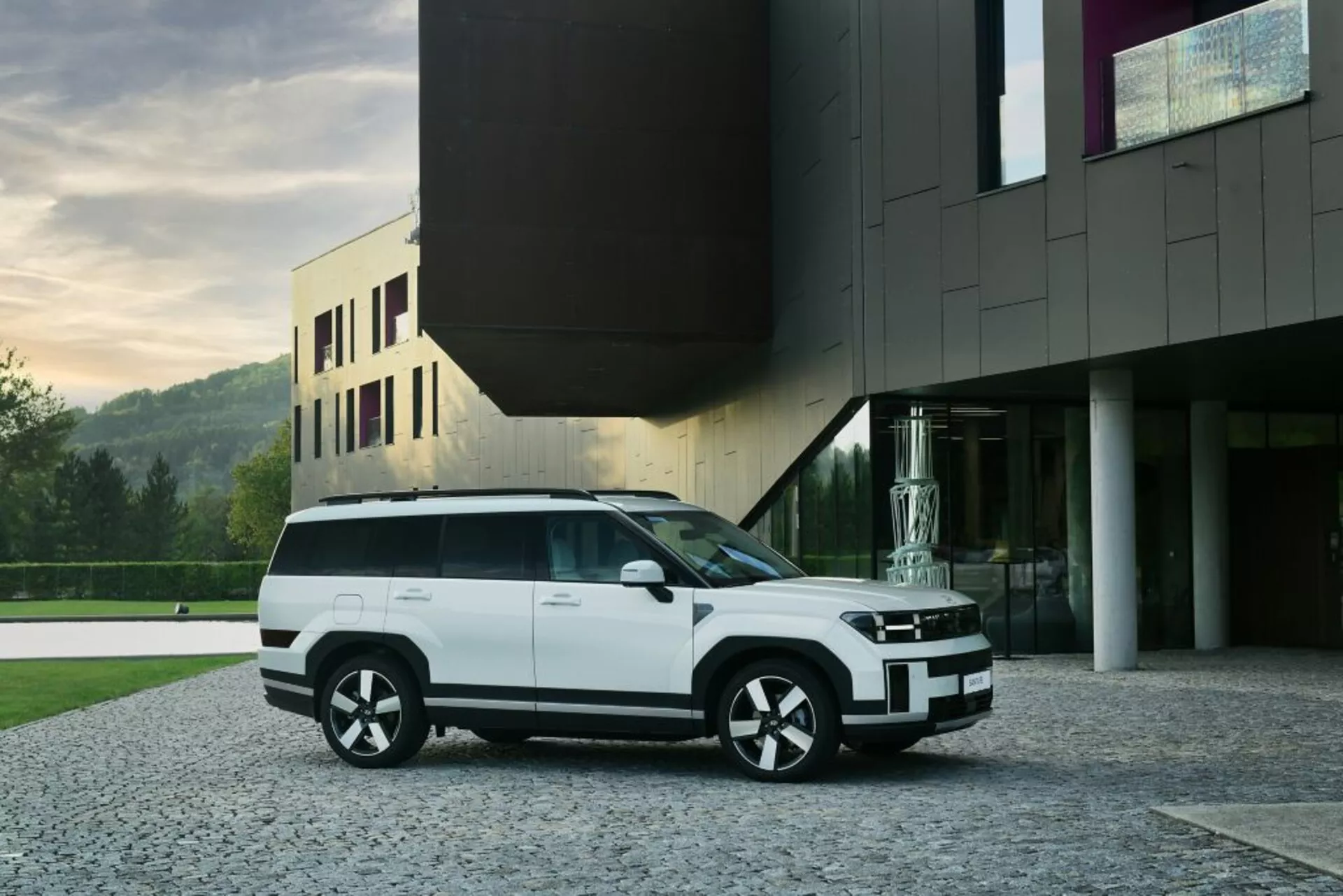 Obraz przedstawia nowoczesny, biały SUV Hyundai Santa Fe 2024 (5. generacja) zaparkowany na brukowanym podjeździe przed współczesnym budynkiem. Uchwycony z perspektywy trzech czwartych z przodu, SUV ukazuje elegancki przedni grill zintegrowany z poziomymi reflektorami, zderzak z czarnymi akcentami i światła przeciwmgielne. Profil boczny ukazuje duże okna z czarnymi obramowaniami, wpuszczane klamki drzwi i mocną linię ramion. Wieloszprychowe koła w dwukolorowym wykończeniu uzupełniają design pojazdu, natomiast płaska linia dachu z ewentualnym panoramicznym szyberdachem i tylnym spojlerem podkreśla jego aerodynamikę. Obraz sugeruje technologię LED w systemie oświetleniowym, podkreślając nowoczesną estetykę pojazdu. Współczesna architektura tła dodaje luksusowego wyglądu SUV.