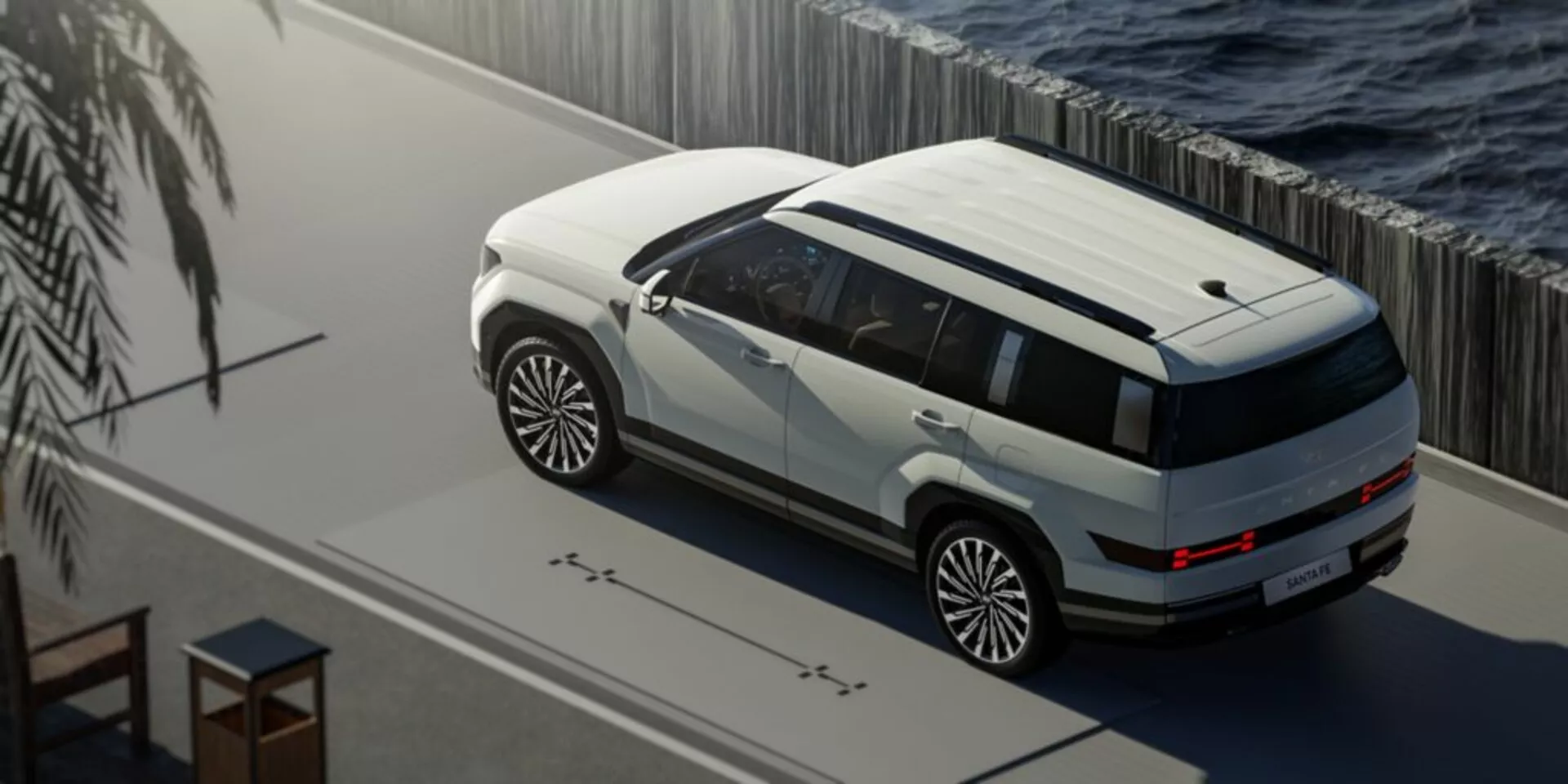 Zdjęcie z wysokiego kąta SUV-a Hyundai Santa Fe piątej generacji z 2024 roku zaparkowanego w pobliżu zbiornika wodnego, z dwukolorowym malowaniem na biało i ciemno. Zdjęcie podkreśla panoramiczny dach, duże wieloramienne felgi aluminiowe, osłony nadwozia i eleganckie oświetlenie LED. Scena jest spokojna, z naturalnym oświetleniem wskazującym na poranek lub popołudnie, i sugeruje luksusowe lub premium otoczenie z potencjalnymi zaawansowanymi funkcjami technologicznymi, takimi jak czujniki zbliżeniowe i czujniki światła otoczenia.