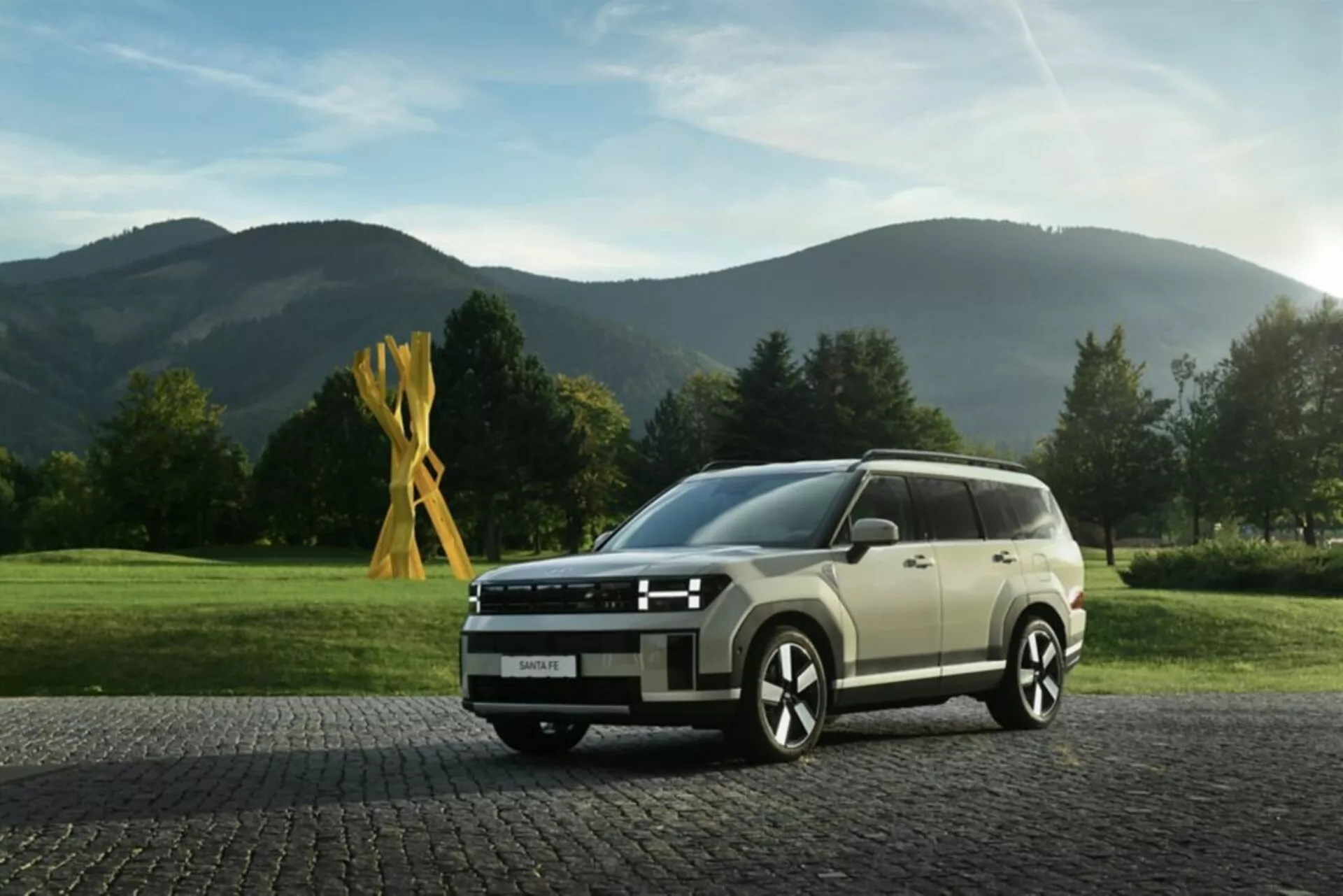 Obraz przedstawia Hyundai Santa Fe 2024, 5. generacji, uchwycony w niskim kącie, z widokiem na przedni kwadrant, który podkreśla nowoczesny i kanciasty design SUV-a. Widoczne cechy to reflektory LED z ostrym sygnaturą świetlną, szeroki przedni grill z poziomymi prętami, częściowo widoczny zderzak z sensorami lub kamerami, wieloszprychowe felgi aluminiowe dla sportowego wyglądu oraz boczne lusterka ze zintegrowanymi kierunkowskazami. Linie karoserii wzdłuż boku podkreślają język projektowania pojazdu, a duże okna mogą oferować filtrację UV. Samochód jest ustawiony na brukowanej nawierzchni z spokojnym, wiejskim tłem, podkreślającym jego wszechstronną atrakcyjność zarówno dla środowisk miejskich, jak i wiejskich. Oświetlenie i głębia ostrości koncentrują uwagę na pojeździe, co czyni go odpowiednim do kategorii takich jak nowoczesne SUV-y, projektowanie samochodów i pojazdy luksusowe.