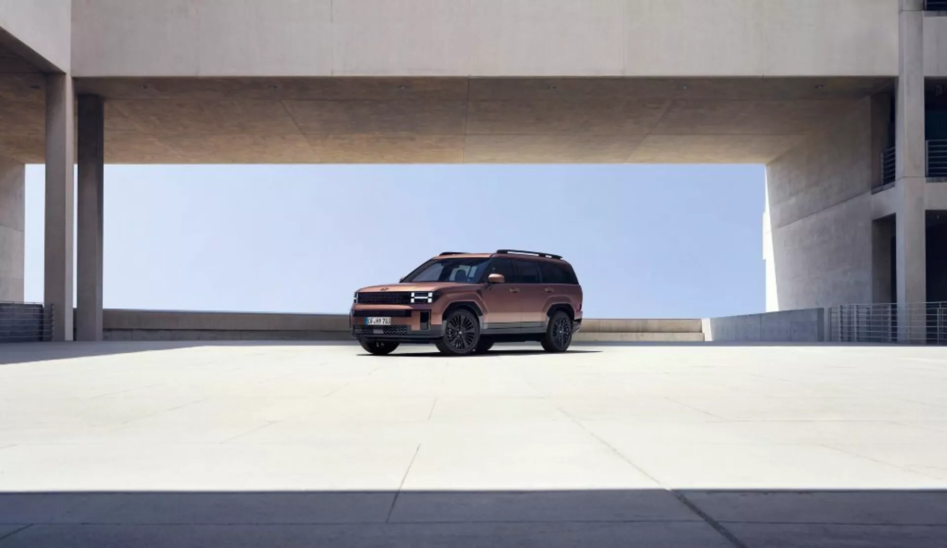 Nowoczesny SUV, prawdopodobnie Hyundai Santa Fe 2024 (5. generacja), jest centralnie umieszczony w przestronnym, minimalistycznym miejskim otoczeniu, prawdopodobnie w betonowej strukturze parkingowej z dużą górną barierą. Uchwycone z średniej odległości, SUV jest pokazany w trójkwartowym widoku z przodu, lekko nachylony w prawo. Samochód charakteryzuje się nowoczesnym designem z eleganckimi liniami, wyrazistym grillem, reflektorami LED, wlotami powietrza, metalowymi felgami, relingami dachowymi, wysokim prześwitem, lusterkami bocznymi z wbudowanymi kierunkowskazami i przyciemnianymi szybami. Naturalne światło oświetla pojazd, podkreślając jego kontury i kolor.