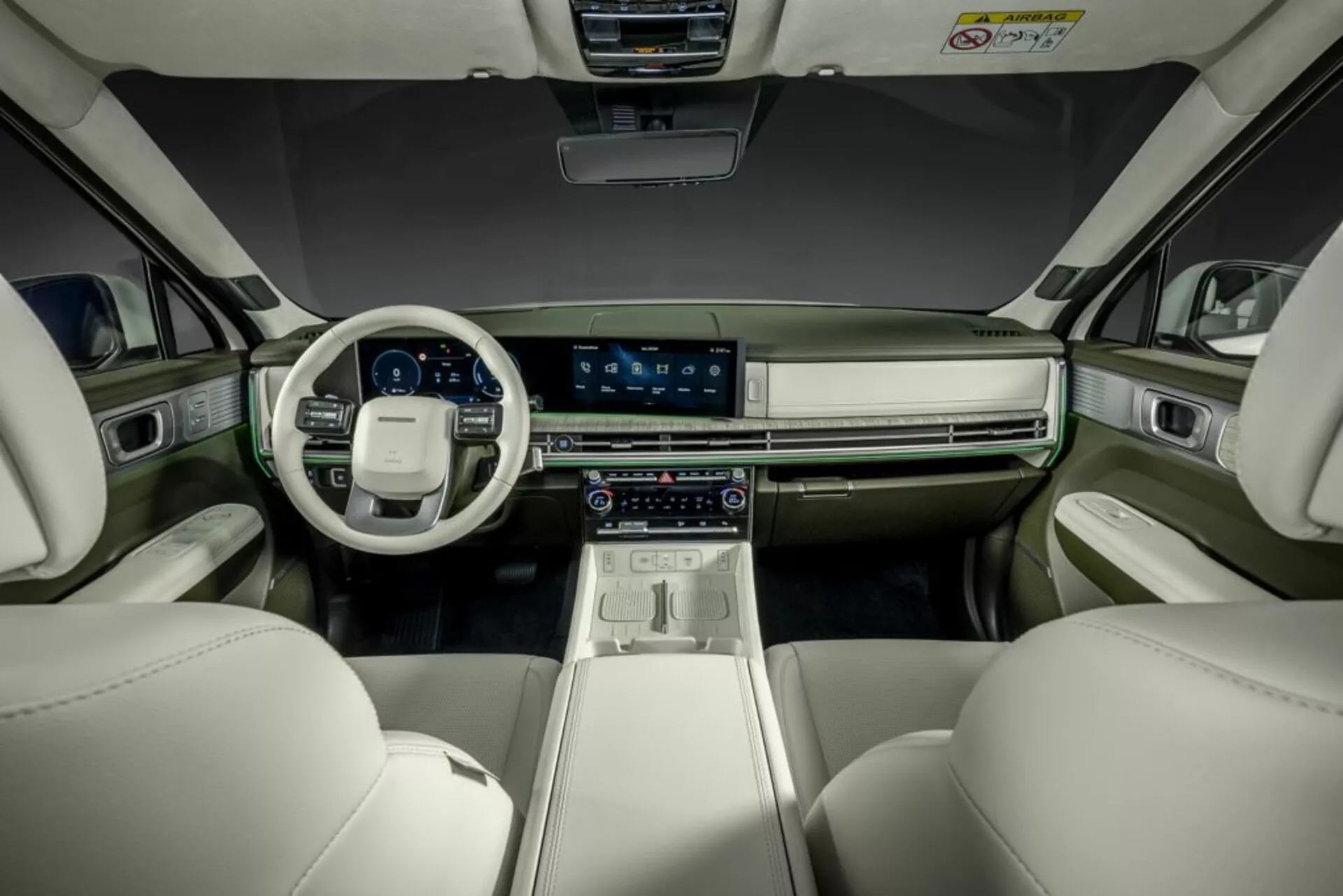 Zdjęcie wnętrza Hyundai Santa Fe 2024 (5. generacji), uchwycone z tylnego siedzenia z widokiem na deskę rozdzielczą. Na zdjęciu widać przednie fotele i kokpit, wyróżniające się dużym, centralnym ekranem dotykowym, cyfrowym zestawem wskaźników, zaawansowanymi kontrolami klimatyzacji oraz kierownicą z zamontowanymi przyciskami sterującymi. Współczesne, luksusowe wnętrze posiada dwukolorowe schematy z jasnymi skórzanymi siedzeniami oraz ciemniejszymi panelami deski rozdzielczej i drzwi, z metalowymi akcentami. Oświetlenie ambientowe poprawia wyrafinowaną atmosferę.