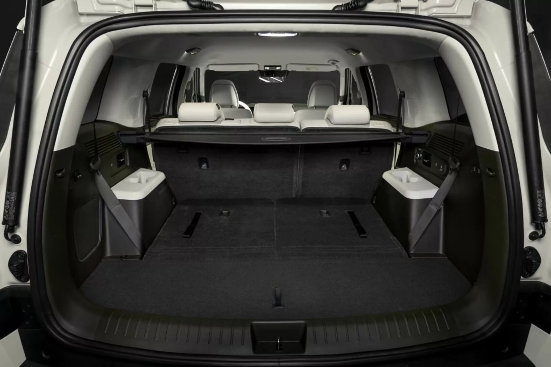 Obraz przedstawia przestrzeń ładunkową wnętrza Hyundai Santa Fe 2024, widzianą z tyłu, z otwartą klapą bagażnika ukazującą płaską podłogę ładunkową pokrytą wykładziną. Trzeci rząd siedzeń jest złożony, aby powiększyć przestrzeń ładunkową, podczas gdy środkowy rząd pozostaje w pozycji pionowej dla pasażerów. Dobrze oświetlone wnętrze podkreśla cechy takie jak haki do mocowania, boczne schowki i zintegrowane panele mogące mieścić głośniki lub kratki wentylacyjne.