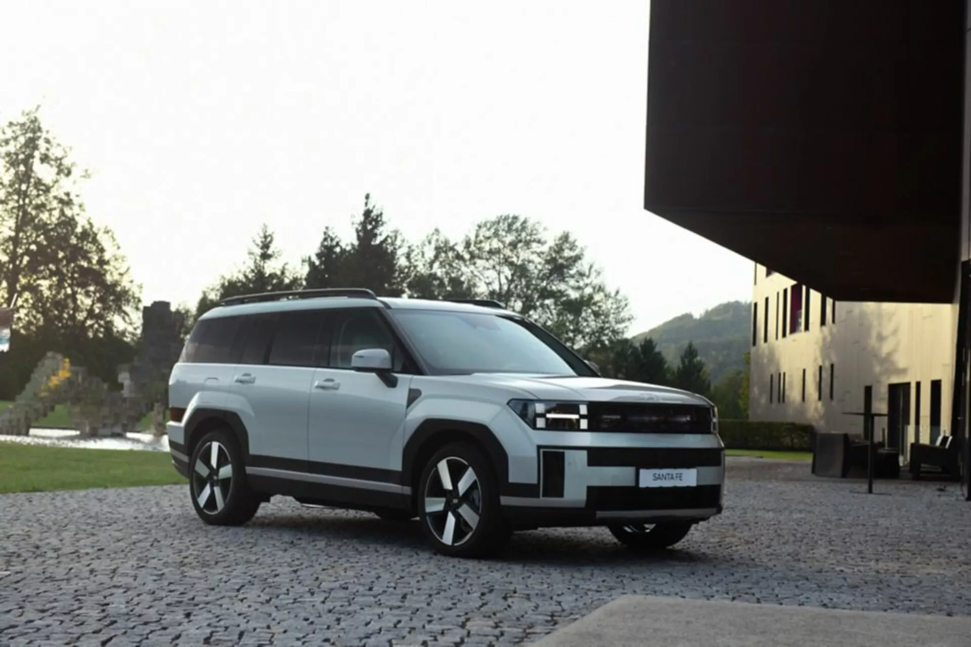Hyundai Santa Fe 2024 piątej generacji w białym kolorze, zaparkowany na brukowanej nawierzchni z nowoczesnym budynkiem i zielenią w tle, widoczny z przedniego lewego boku pod kątem trzech czwartych, z prostokątną przednią kratką, reflektorami LED lub HID oraz felgami aluminiowymi, sfotografowany przy miękkim naturalnym oświetleniu podkreślającym nowoczesną estetykę SUV-a.