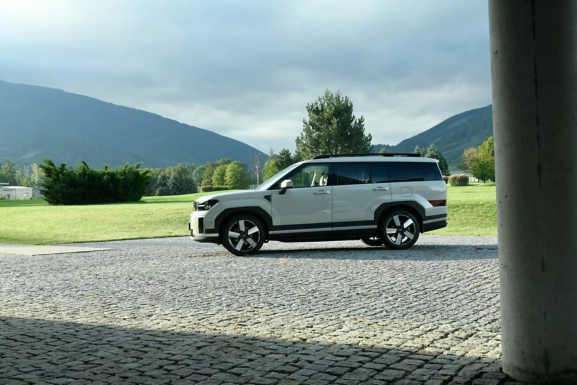 Widok z profilu bocznego SUV-a Hyundai Santa Fe 2024 (5. generacji) zaparkowanego na utwardzonej powierzchni z naturalnym tłem. Obraz uwydatnia nowoczesny design pojazdu, zwracając uwagę na duże felgi z wieloma szprychami, dwukolorowe malowanie i szczegółowe linie nadwozia. Kadr, otoczony kolumną struktury, został uchwycony z niższego kąta, podkreślając wysokość i postawę samochodu. Dzienny plenerowy scenariusz podkreśla zewnętrzne cechy pojazdu bez mocnych cieni.