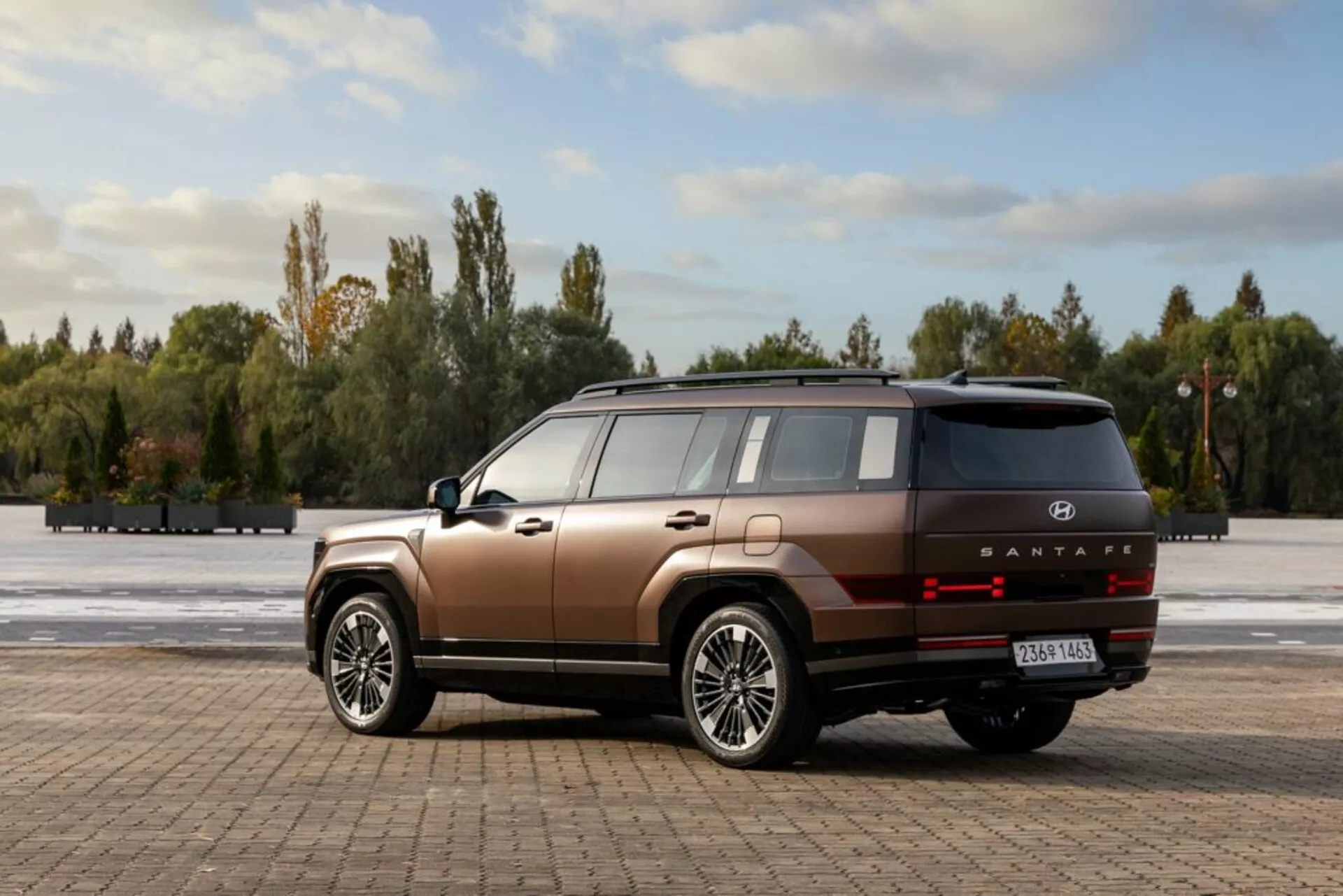 Obraz przedstawia nowoczesny SUV Hyundai Santa Fe 2024, 5. generacji, na otwartej wybrukowanej przestrzeni, otoczony drzewami i niebieskim niebem z rozproszonymi chmurami. Fotografowany pod kątem trzech czwartych od tyłu, pojazd ukazuje wysoką linię boczną, płaskie panele boczne i kanciastych nadkola z dużymi, wieloszprychowymi felgami aluminiowymi. SUV ma dwukolorowe malowanie w miedzianobrązowym kolorze z czarnymi dolnymi listwami. Tylna część zawiera poziomo zorientowane światła tylne LED, srebrny pasek ozdobny, oznaczenie modelu 'SANTAFE' i odblaski na zderzaku. Antena w kształcie płetwy rekina i panoramiczny dach sugerują zaawansowane funkcje technologiczne. Naturalne światło dzienne podkreślające kontury samochodu dopełnia scenę.