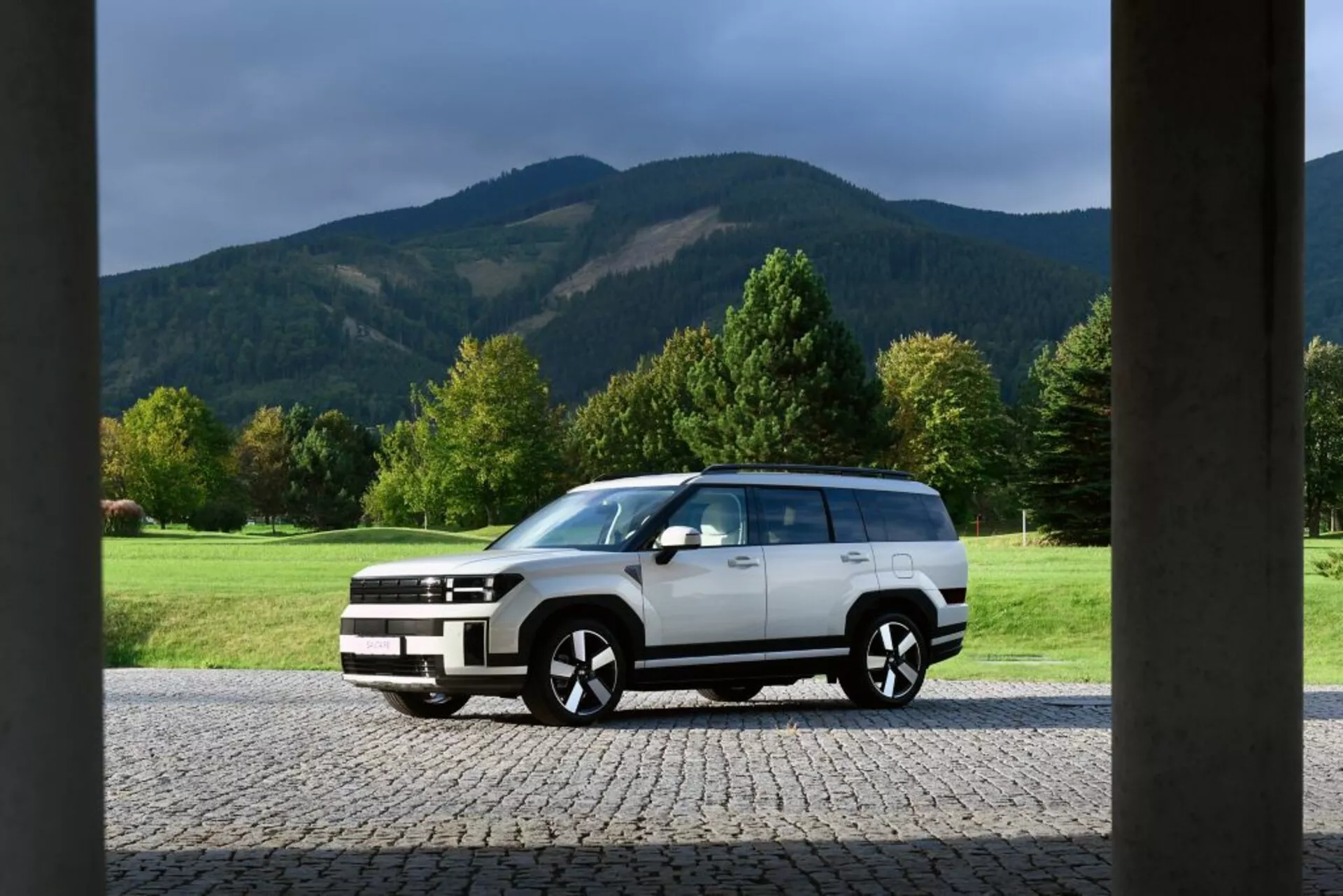 Zdjęcie przedstawia Hyundai Santa Fe 2024, piątą generację, zaparkowaną przed budynkiem z dużymi kolumnowymi strukturami na brzegach. Samochód to pełnowymiarowy SUV o pudełkowym, muskularnym designie, przedstawiający nowoczesną estetykę siły i solidności. Ma dwukolorowe malowanie z białym nadwoziem i czarnymi wykończeniami na dolnej części i nadkolach. SUV wyposażony jest w duże aluminiowe felgi, przyciemniane szyby, reflektory LED, światła dzienne i relingi dachowe, co świadczy o jego zaawansowanym designie i cechach ukierunkowanych na użytkowość. Sceniczny krajobraz w tle obejmuje otwarte trawiaste pole, drzewa i góry, podkreślając przygodowe środowisko na świeżym powietrzu.