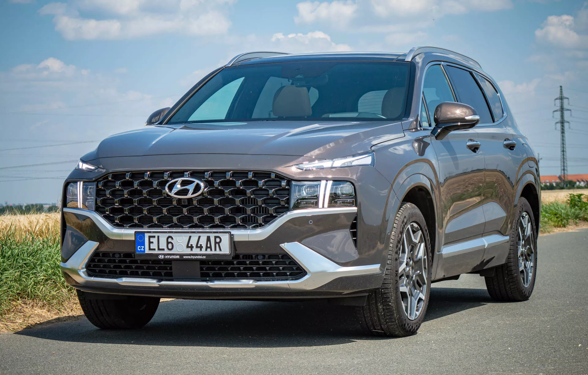Obraz przedstawia SUV Hyundai Santa Fe 2020 zaparkowany na asfaltowej drodze, sfotografowany z przedniego lewego trzyczwrtowego kąta. Wyraźnie pokazuje dużą kratkę z charakterystycznym wzorem marki i centralnym logo, stylowe reflektory LED i wyrzeźbioną maskę. Szeroka przednia szyba zapewnia dobrą widoczność, a boczny profil ukazuje wieloramienne felgi aluminiowe i lusterko boczne kierowcy z potencjalnie zintegrowanymi kierunkowskazami. Ciemny kolor pojazdu podkreśla jego wyrafinowany wygląd z nowoczesnymi elementami designu, w tym możliwymi czujnikami lub kamerami przednimi.