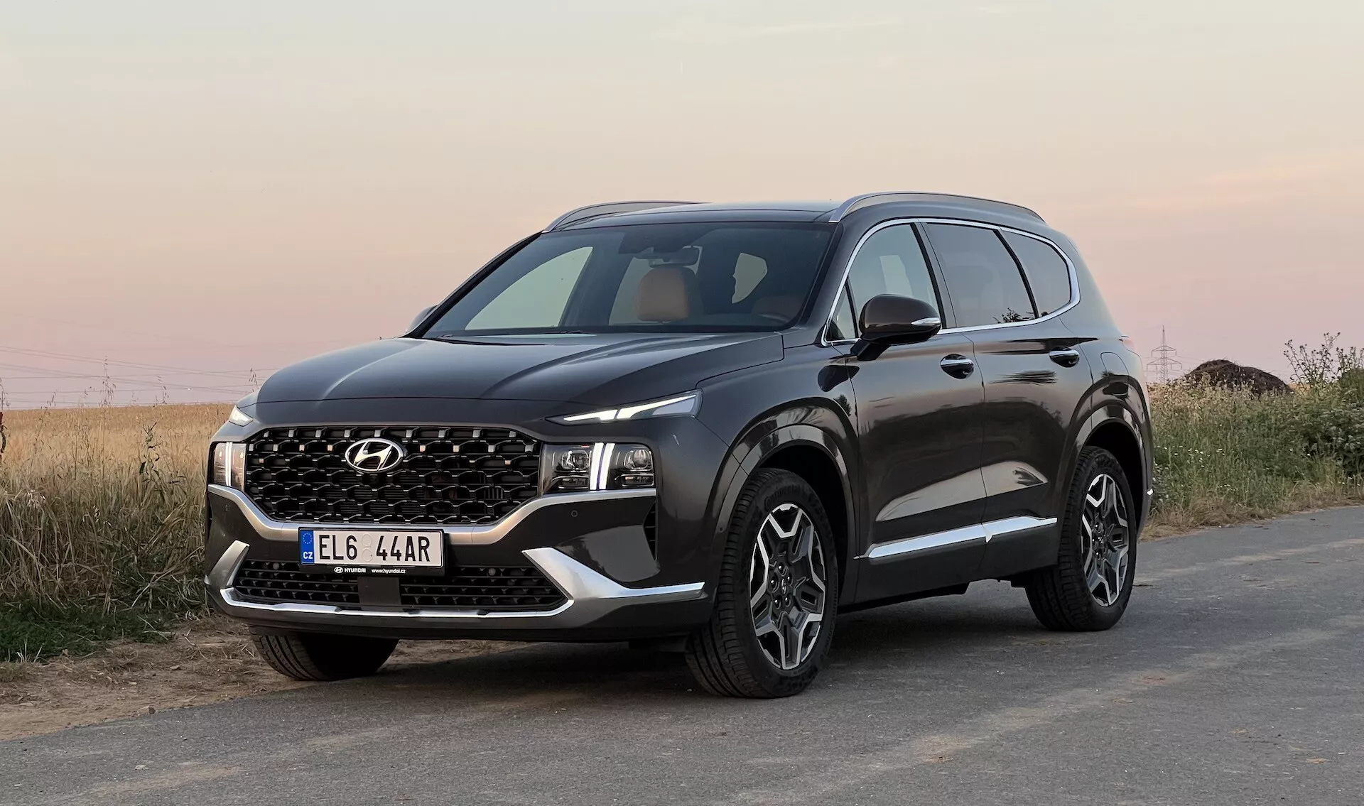 Obraz SUV Hyundai Santa Fe 2020 zaparkowanego na powierzchni asfaltowej, wykonany z niskiego kąta z przednim widokiem trzy-czwartym, pokazujący przedni grill, wąskie reflektory LED, zderzak z światłami przeciwmgielnymi i fałszywą płytą poślizgową, maskę z ostrymi zagnieceniami, duże wieloramienne felgi aluminiowe, czarne osłony ochronne na nadkolach i dolnej części drzwi, boczne lusterko z zintegrowanymi kierunkowskazami oraz okna z chromowanymi ramkami. Tło przedstawia nieokreślony naturalny krajobraz pod otwartym niebem.