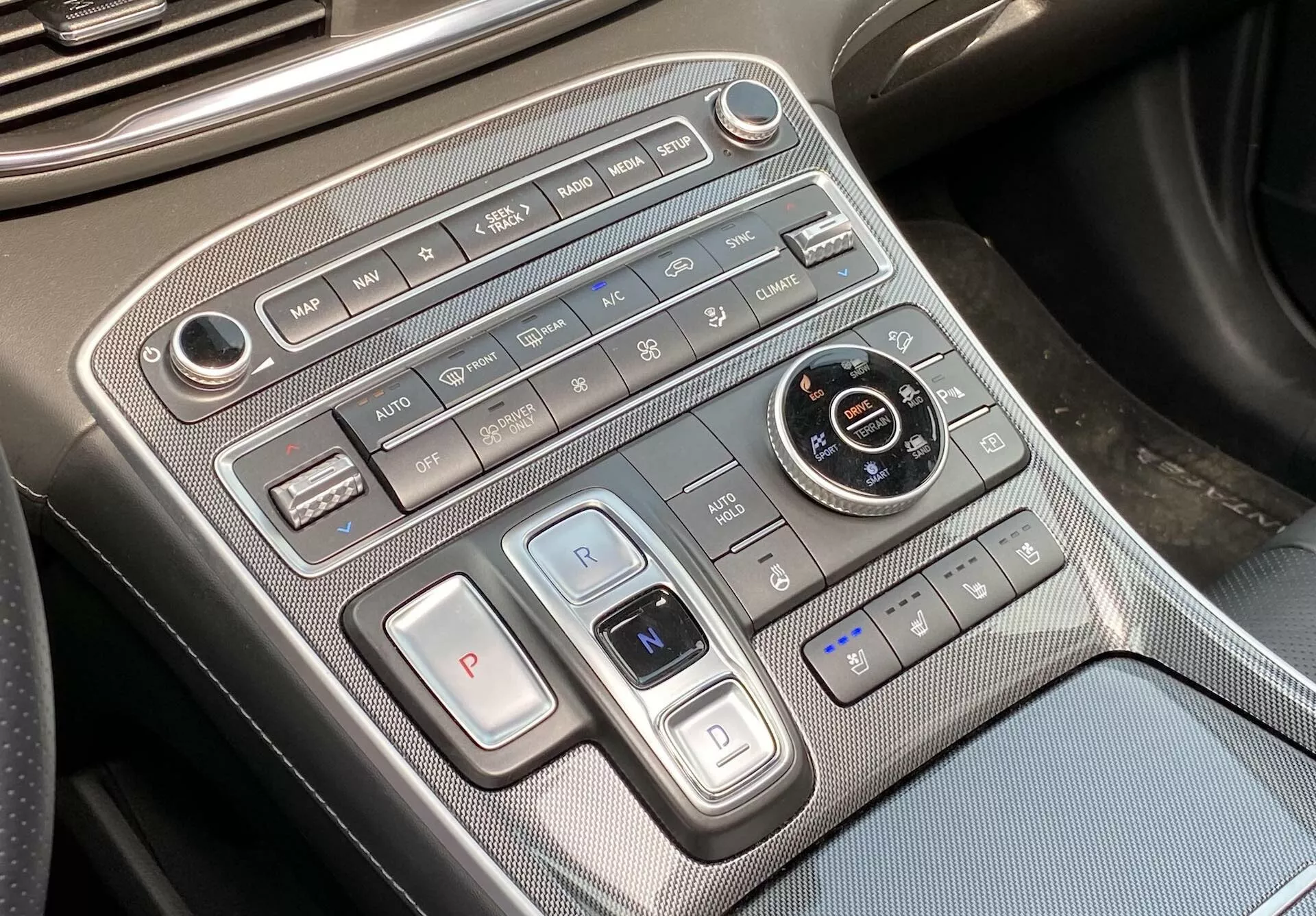 Na tym zdjęciu przedstawiono zbliżenie konsoli centralnej w Hyundai Santa Fe 2020, skupiając się na obszarze sterowania klimatyzacją i przekładnią. Ujęcie, lekko nachylone z perspektywy kierowcy lub pasażera, odsłania umieszczony poziomo panel sterowania klimatyzacją na górze, zawierający kontrolki prędkości wentylatora, przyciski przepływu powietrza, automatyczną klimatyzację, odmrażacz i recyrkulację powietrza. Niżej znajdują się przyciski wyboru biegów z dużym przyciskiem „P” do parkowania i przełącznikiem przypominającym dźwignię do biegów „R” i „N”, co wskazuje na użycie elektronicznego lub przyciskowego systemu wyboru biegów. Okrągłe pokrętło do wyboru trybów jazdy, otoczone przyciskami hamulca auto-hold, wspomagania parkowania i systemem automatycznego start/stop, jest obecne. Kontrolki foteli podgrzewanych lub wentylowanych są widoczne w prawym dolnym rogu. Konsola wyróżnia się wysokiej jakości materiałami z metalowymi akcentami i wzorem przypominającym włókno węglowe. Widoczny jest port USB w lewym dolnym rogu, oferujący opcje łączności z urządzeniami.