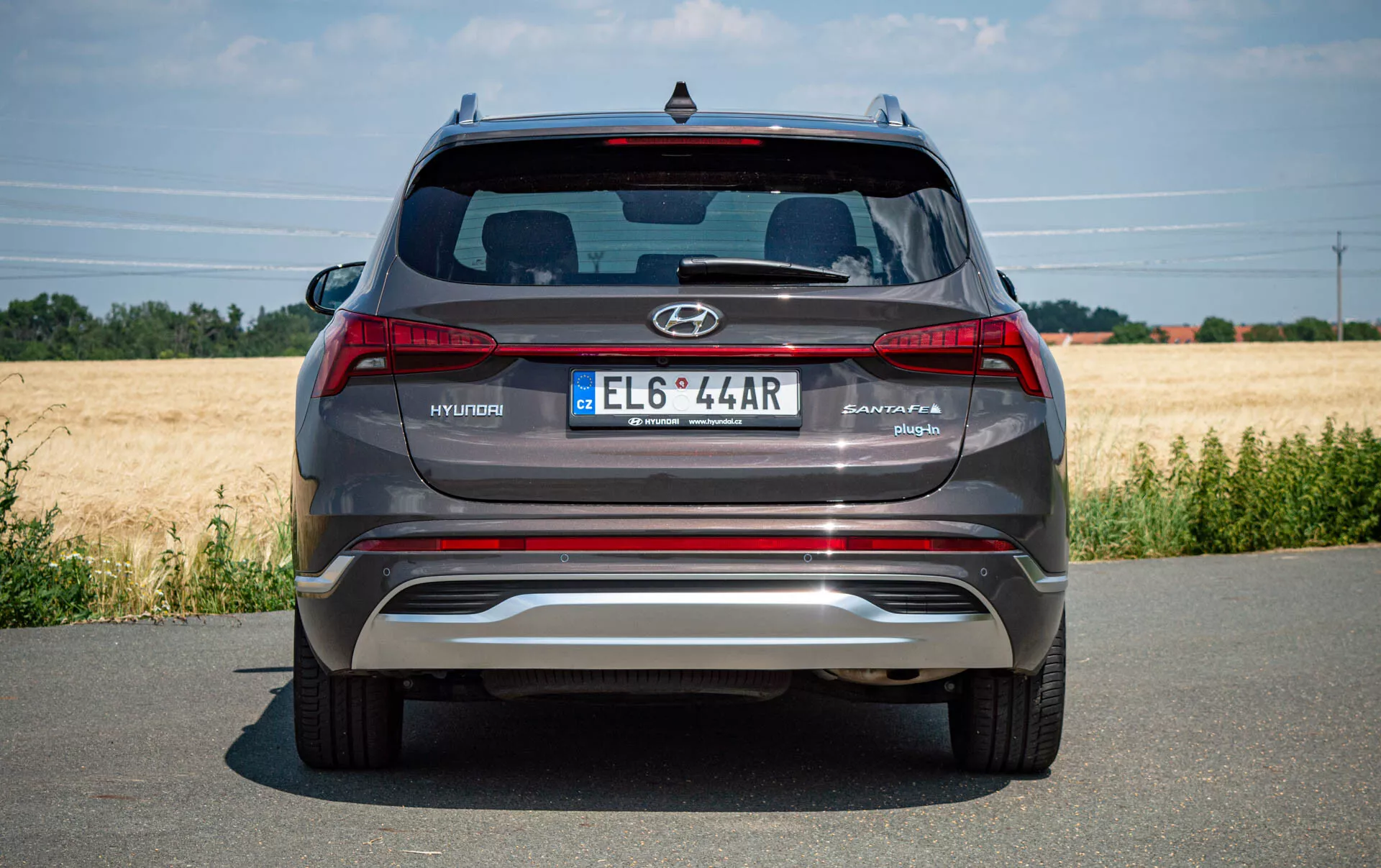 Tylny widok nowoczesnego szaro-srebrnego SUVa Hyundai Santa Fe 2020 zaparkowanego na brukowanej drodze, z logo Hyundaia, nazwą modelu Santa Fe, europejską tablicą rejestracyjną, tylnymi światłami, odblaskowym metalowym tylnym zderzakiem z wbudowanymi czujnikami parkowania, podwójnymi końcówkami wydechu, tylną szybą ze spojlerem Hyundai i trzecim światłem stopu. Czysta pogoda z polem w tle.