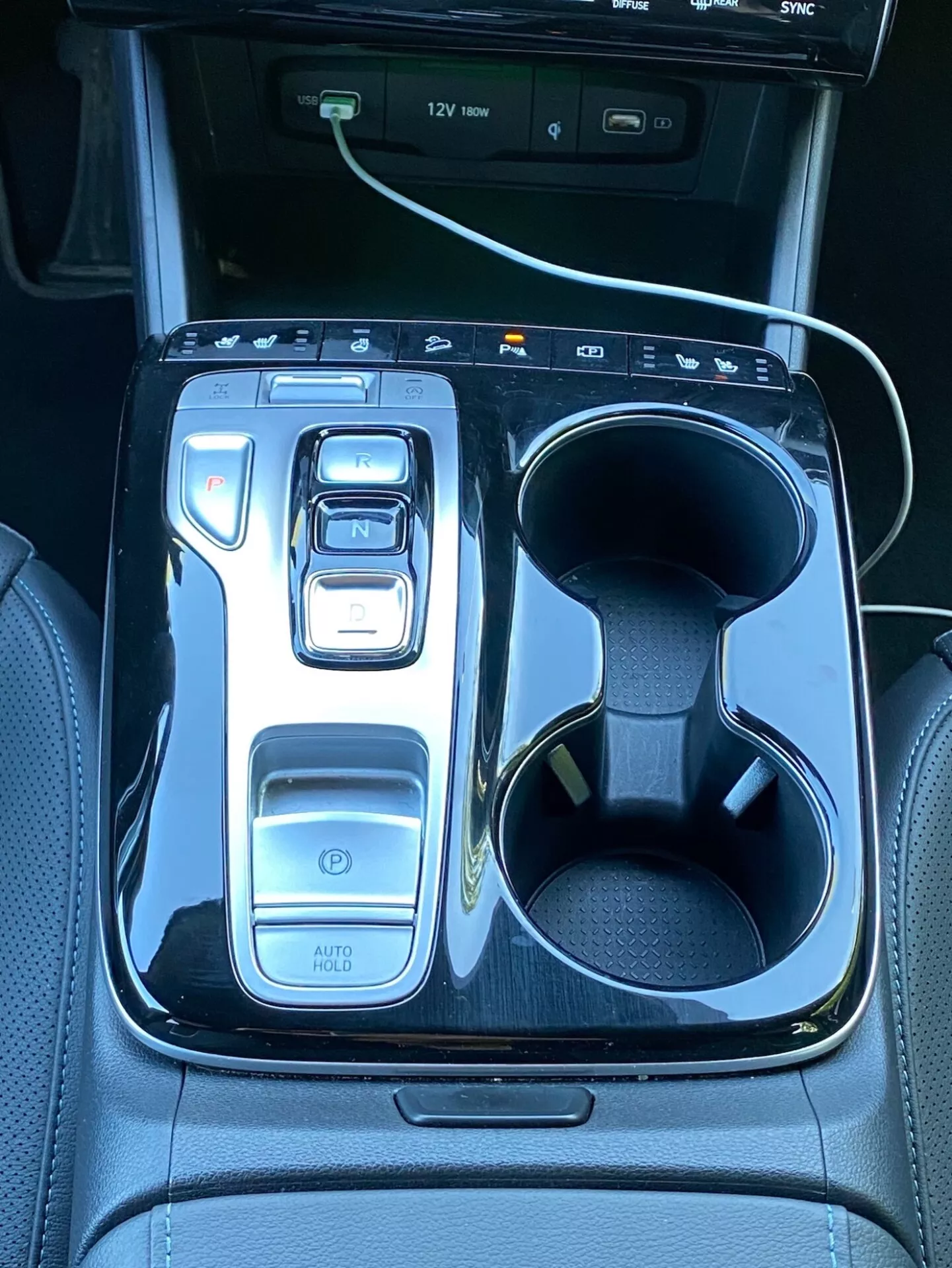 Obraz przedstawia centralną konsolę Hyundai Tucson 2020, z elektronicznym selektorem biegów ustawionym na 'N' otoczonym błyszczącą czarną wykończeniem. Po lewej stronie znajdują się przyciski, w tym przycisk włączania, wybieraki trybów jazdy oraz funkcja 'AUTO HOLD'. Po prawej stronie umieszczono podwójne uchwyty na kubki, z jednym z nich zajętym przez biały kabel ładujący. W górnej części widać fragment ekranu infotainment z ikonami portów USB, podkreślającą technologię i luksus pojazdu.