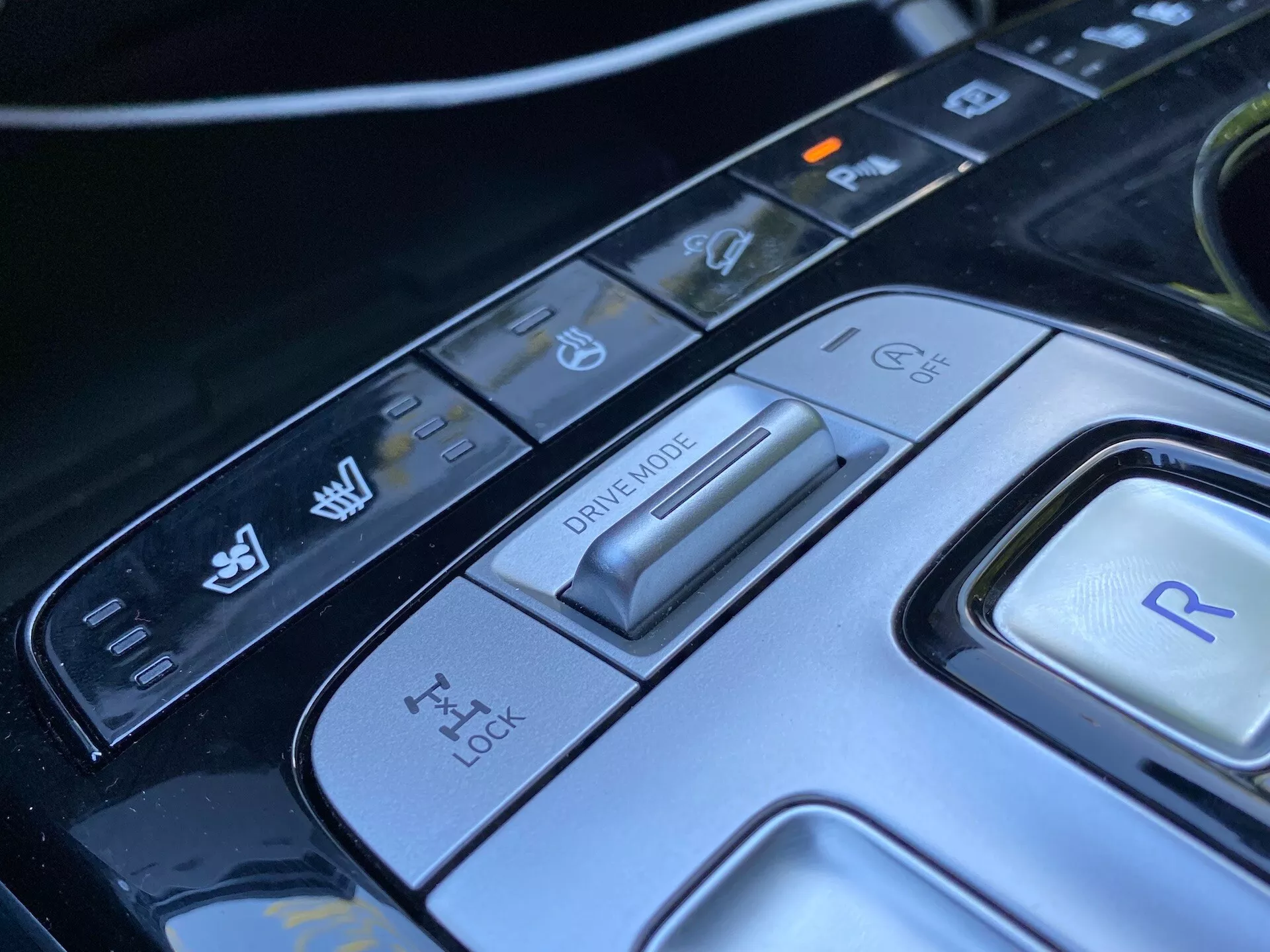 Zbliżony widok konsoli centralnej Hyundai Tucson 2020, skoncentrowany na selektorze trybu jazdy i różnych przyciskach sterowania. Obraz pokazuje zaawansowany przełącznik trybu jazdy na różne warunki jazdy, panel kontroli trakcji, przycisk do wstecznego biegu i dodatkowe kontrolki stabilności pojazdu oraz funkcje wspomagania parkowania. Elegancki design i nowoczesne materiały podkreślają wyrafinowane wnętrze samochodu tego luksusowego, nowoczesnego samochodu.