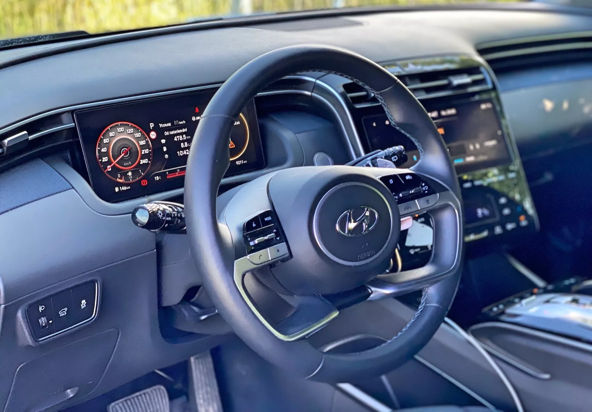 Wnętrze samochodu Hyundai Tucson 2020 z perspektywy kierowcy, z nowoczesną kierownicą z wielofunkcyjnymi przyciskami i cyfrowym panelem instrumentów wyświetlającym prędkościomierz, obrotomierz, poziom paliwa i temperaturę silnika. Zdjęcie podkreśla również część konsoli środkowej z dużym ekranem dotykowym systemu infotainment, nowoczesny design deski rozdzielczej z oświetleniem ambientowym i materiały wysokiej jakości sugerujące luksusowe wykończenia.