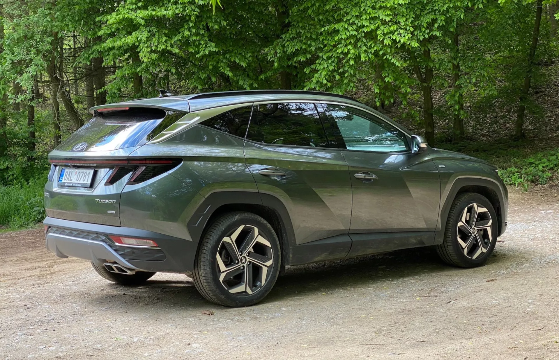Tylny widok trzech czwartych nowoczesnego SUV-a Hyundai Tucson 2020 z naciskiem na boczny profil i tylny design. Pojazd jest ustawiony po lewej stronie, zdjęcie wykonane z prawej, podkreślając tylny prawy róg. Charakterystyczne tylne światła LED owijają się od tyłu do boków, a zamontowany na dachu spojler dodaje sportowego akcentu. Wznosząca się linia boczna, opadająca linia dachu, przyciemniane szyby i klamki o kolorze nadwozia podkreślają jego wygląd typu coupé. Duże felgi aluminiowe z wieloramiennym wzorem i niskoprofilowe opony wzmacniają luksusowy wygląd. Drobne detale, takie jak tylna wycieraczka i osłony nadkoli, oznaczają praktyczność i solidność. Samochód jest umieszczony na żwirze w naturalnym otoczeniu z drzewami w tle.