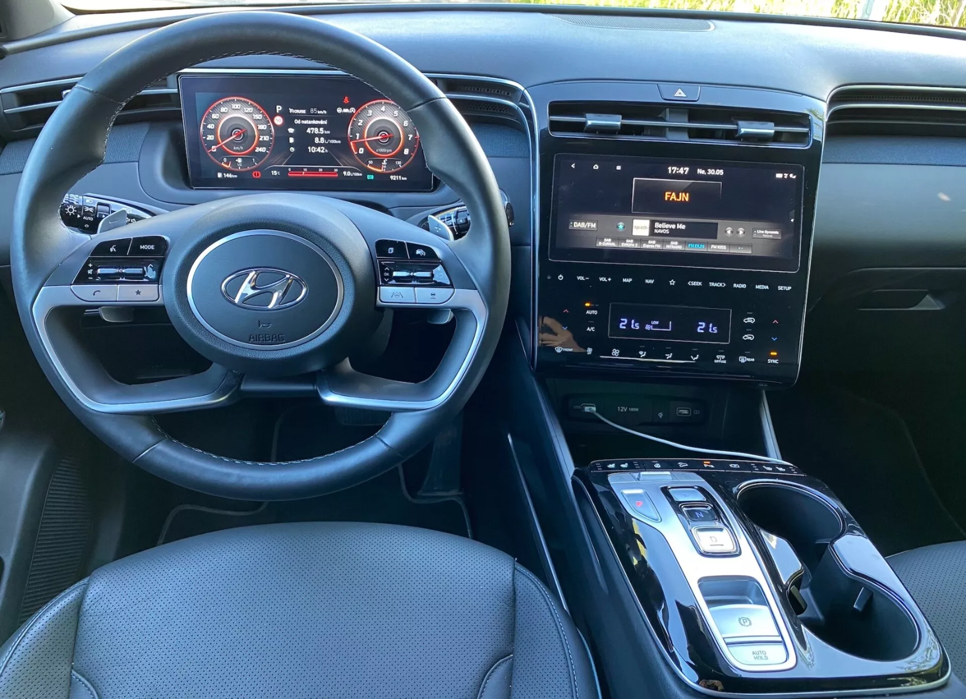 Widok wnętrza Hyundai Tucson 2020 z fotela kierowcy, ukazujący kierownicę z logo Hyundai oraz przyciski do sterowania audio, telefonem i tempomatem. Deska rozdzielcza posiada cyfrowy panel instrumentów z prędkościomierzem i obrotomierzem z czerwonymi akcentami. Konsola centralna wyposażona jest w system infotainment z ekranem dotykowym wyświetlającym sterowanie klimatyzacją, fizycznymi przyciskami i pokrętłami oraz automatyczną dźwignię zmiany biegów z elektronicznym selektorem. Widoczna jest potencjalna bezprzewodowa ładowarka, ciemna tapicerka materiałowa i dobrze oświetlona kabina.