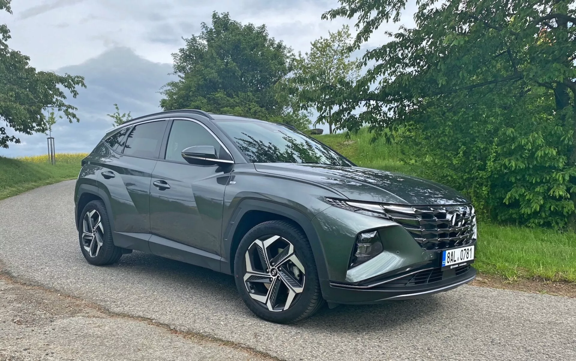 Obraz przedstawia nowoczesny SUV Hyundai Tucson 1.6 T-GDi Pure zaparkowany na asfaltowej drodze z naturalnym krajobrazem w tle. Fotografowany pod przednim kątem trzech czwartych, widoczne są wyraźny grill, eleganckie reflektory, przedni zderzak z światłami przeciwmgielnymi lub wlotami powietrza, duże aluminiowe felgi o wieloramiennym wzorze oraz lusterka boczne. Sylwetka SUV-a opada w kierunku tyłu, co wskazuje na sportowy, aerodynamiczny design. Uchwycona w miękkim świetle dziennym, fotografia podkreśla kolor i detale pojazdu bez ostrych cieni.