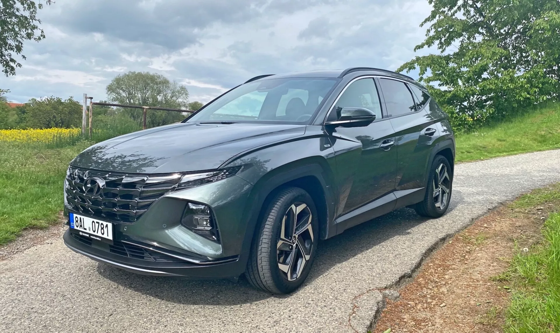 Hyundai Tucson 2020 1.6 T-GDI Pure SUV, widziany z przedniego trójkwartalnego kąta na asfaltowej drodze z drzewami i trawą w tle. Obraz ukazuje grille samochodu, ostre reflektory LED, aluminiowe felgi, lusterka boczne z wbudowanymi kierunkowskazami i ciemnym metalizowanym lakierem. Widoczne są przednie drzwi kierowcy i tylne. Zdjęcie zrobiono w ciągu dnia, przy pochmurnej pogodzie.