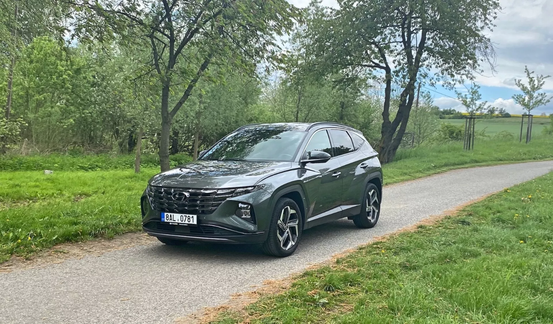 Trzyczwarte widok z przodu crossovera SUV Hyundai Tucson 2020 zaparkowanego na brukowanej ścieżce z zielenią w tle, ukazujący charakterystyczną przednią kratkę pojazdu, nowoczesne reflektory LED, duże aluminiowe koła wieloramienne, opadającą linię dachu, wysoki prześwit, zintegrowane lusterka boczne, przyciemniane szybki i stylowe kontury nadwozia.