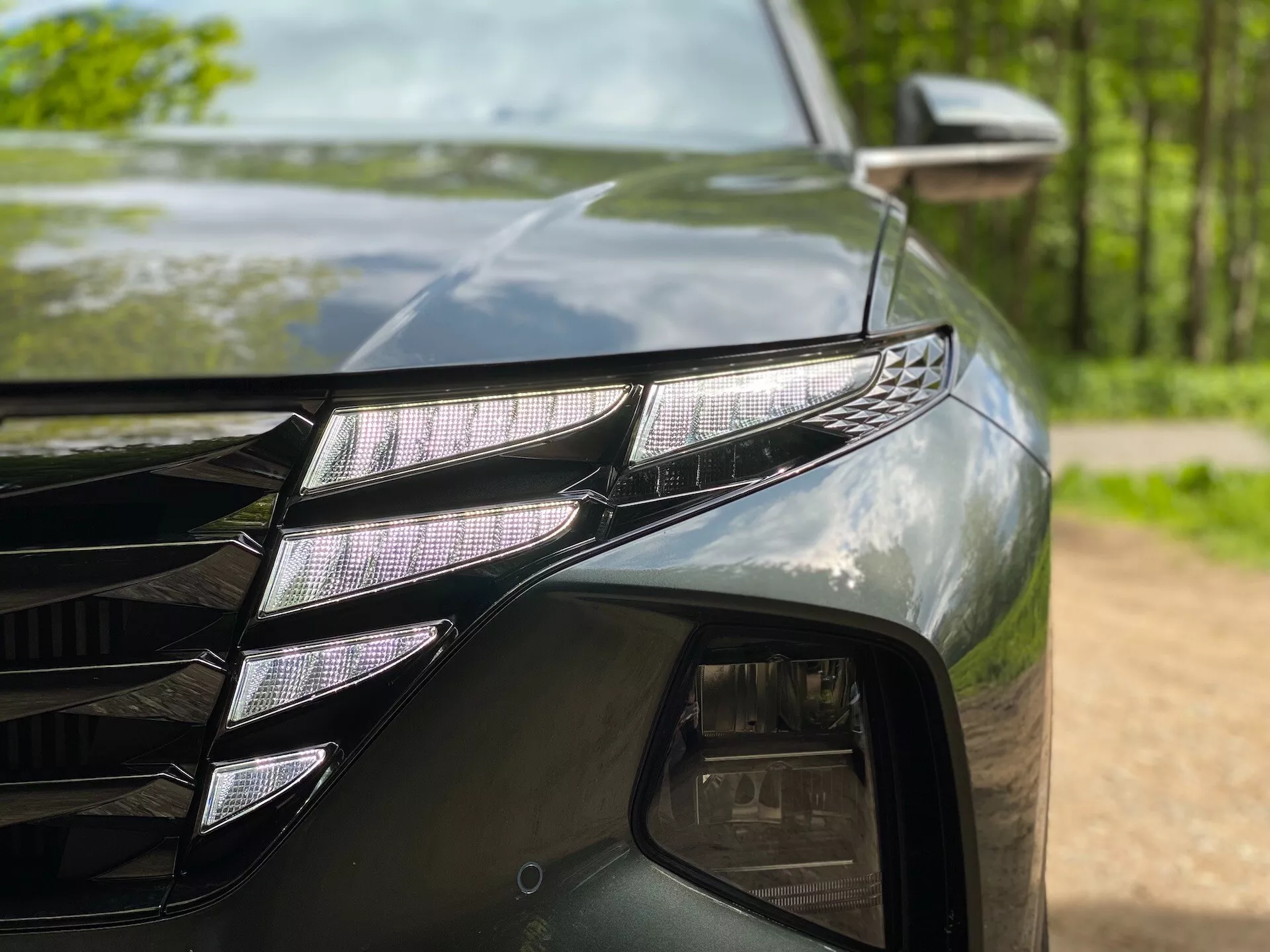 Zbliżenie przedniej lewej części Hyundai Tucson 2020 (Hyundai-Tucson-1-6-T-GDi-Pure), ukazującej zestaw lamp LED z indywidualnymi projektorami LED i charakterystycznym paskiem LED DRL. Ciemny, błyszczący lakier odbija otaczające drzewa. Widoczna jest również część kratki z kanciastymi liniami oraz lusterko boczne z wskaźnikiem kierunkowskazu.