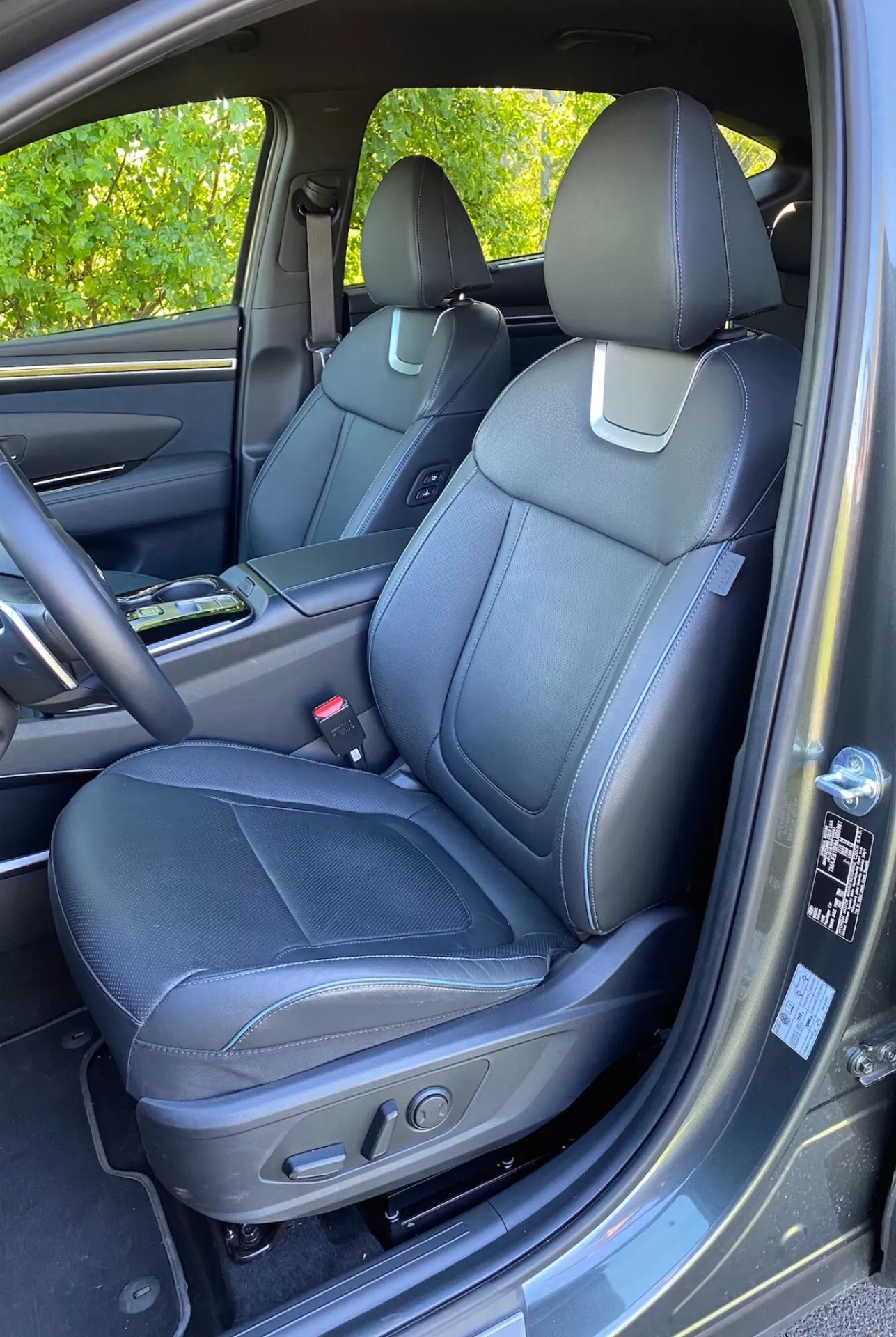 Wnętrze 2020 Hyundai Tucson 1.6 T-GDI Pure ukazujące przednie siedzenie pasażera z czarną skórzaną tapicerką, szczegółowymi szwami i elektrycznymi kontrolkami regulacji. Widoczna środkowa konsola z uchwytem na kubki, panel drzwi z dopasowanymi materiałami, uchwyt, schowek i kratka głośnika sugerująca zaawansowany system audio. Widoczne częściowo deska rozdzielcza i schowek. Dywanik przypominający dywan i delikatne odbicie światła słonecznego na panelu drzwi.