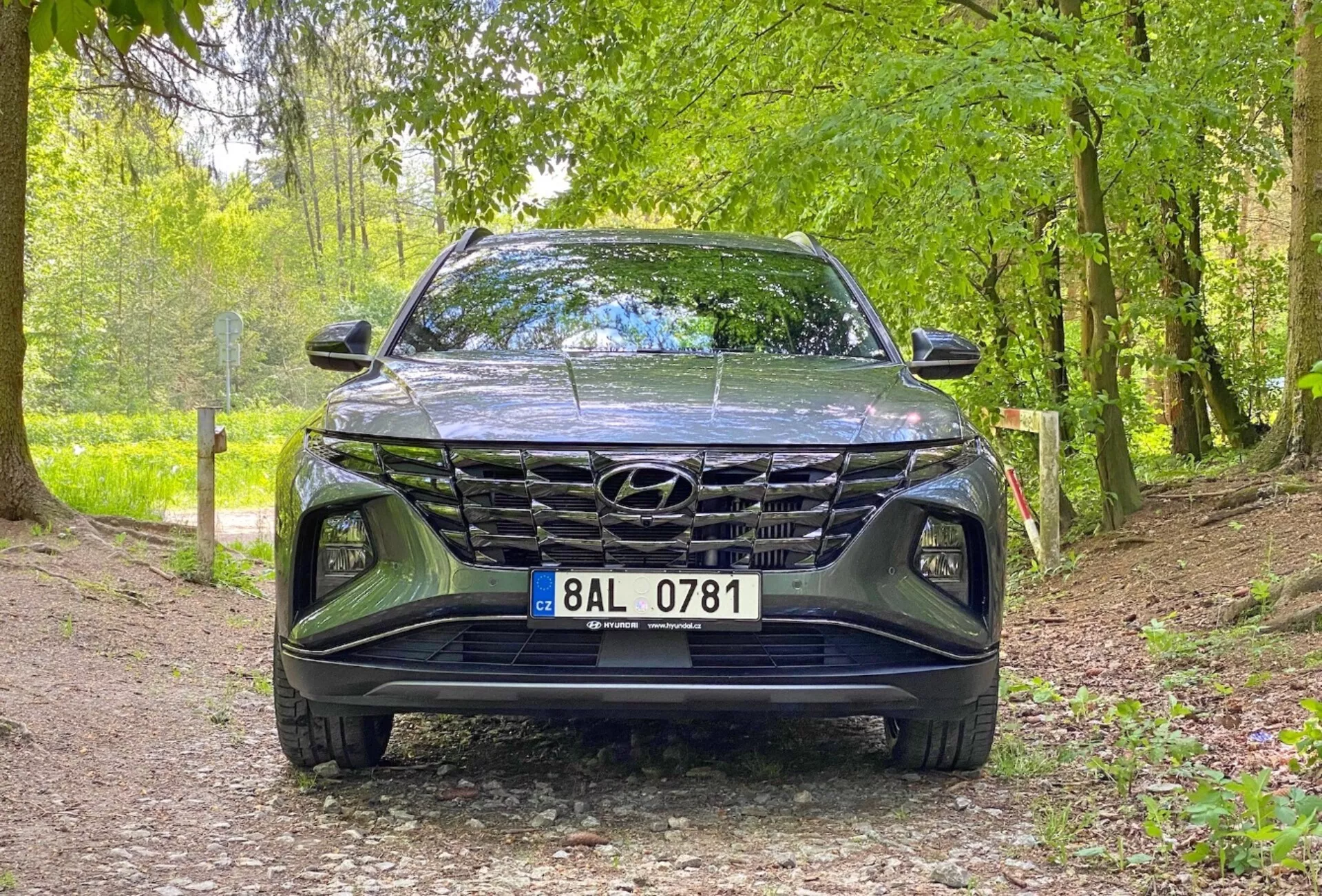Widok z przodu pojazdu Hyundai Tucson 1.6 T-GDi Pure 2020, pokazujący ujęcie z niskiego kąta, skupione na grillu, przednim zderzaku i obszarze maski. Samochód zaparkowany jest na żwirowej ścieżce w naturalnym otoczeniu, co sugeruje jego przydatność w środowiskach zewnętrznych. Kluczowe cechy to: wyraźny design grilla, smukłe reflektory LED i widoczne logo Hyundai.