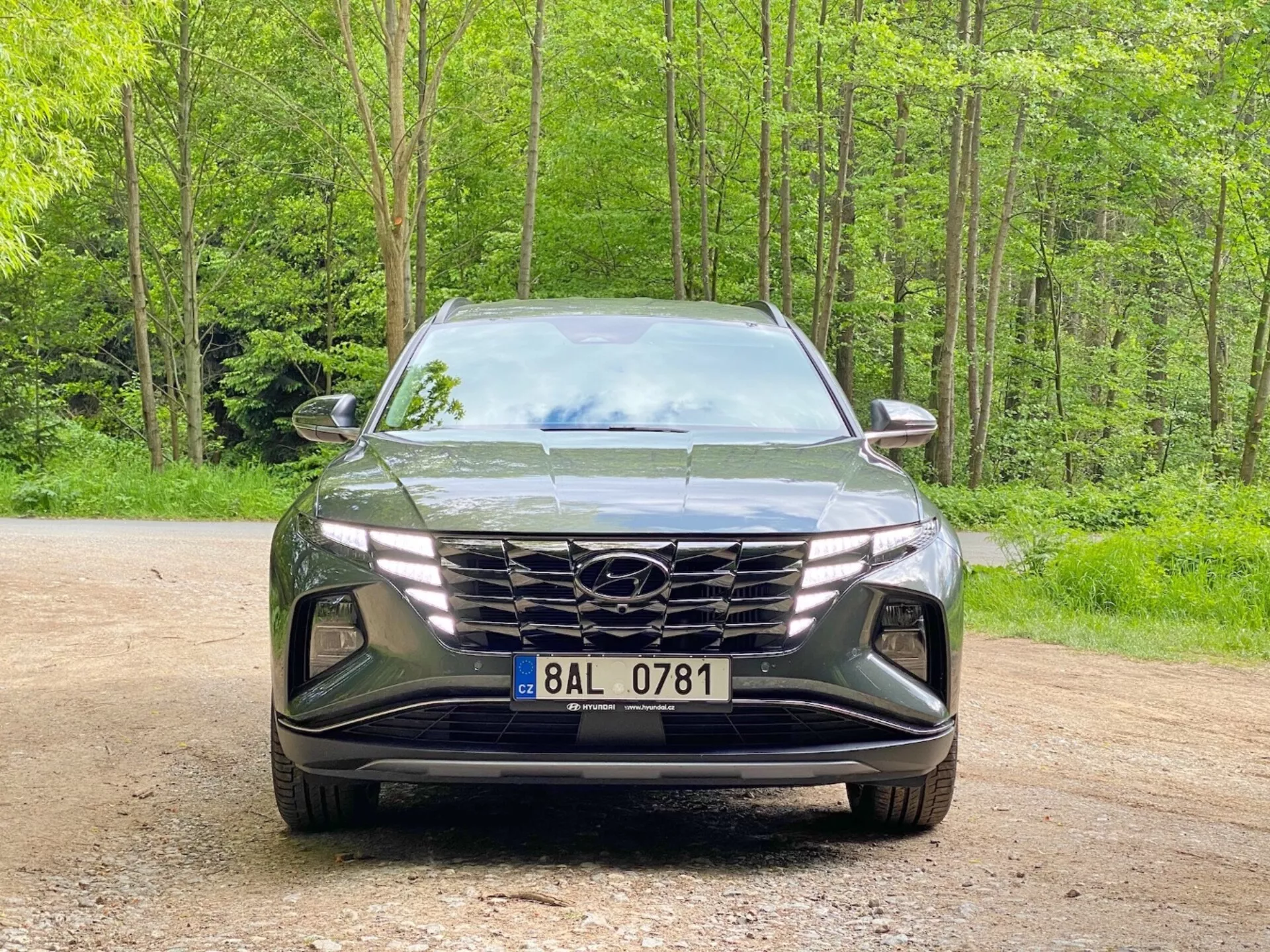 Widok z przodu Hyundai Tucson 2020 (Hyundai Tucson 1.6 T-GDI Pure) nowoczesnego SUV-a zaparkowanego na żwirowej ścieżce z leśnym tłem. Obraz przedstawia elegancki, agresywny design z dużym geometrycznym grillem z logo producenta, wąskimi, kątowymi reflektorami LED i rzeźbionym przednim zderzakiem z wlotami powietrza i wnękami na reflektory przeciwmgielne, podkreślając zaawansowaną aerodynamikę i technologię oświetleniową.
