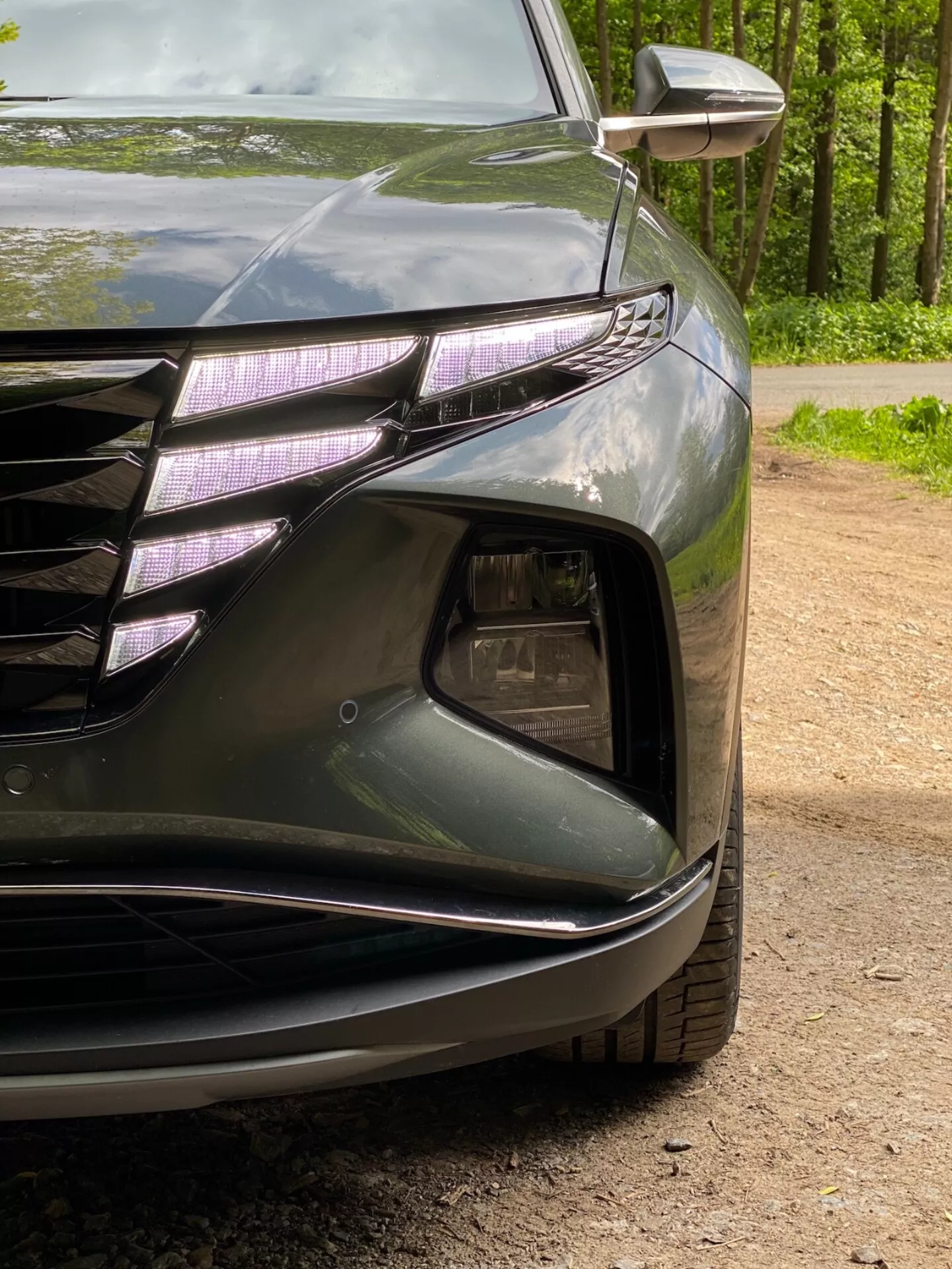 Zbliżenie na lewy przedni róg Hyundai Tucson 2020 1.6 T-GDI Pure, ukazujące reflektor z diodami LED do jazdy dziennej i soczewką typu projektor, część przedniego grilla z poziomymi liniami i chromowanymi akcentami, sekcję wlotu powietrza z obudową lamp przeciwmgielnych, rzeźbiony przedni zderzak z gładkimi liniami oraz zintegrowany kierunkowskaz w lusterku zewnętrznym. Ciemny błyszczący lakier podkreśla aerodynamiczne krzywizny samochodu.