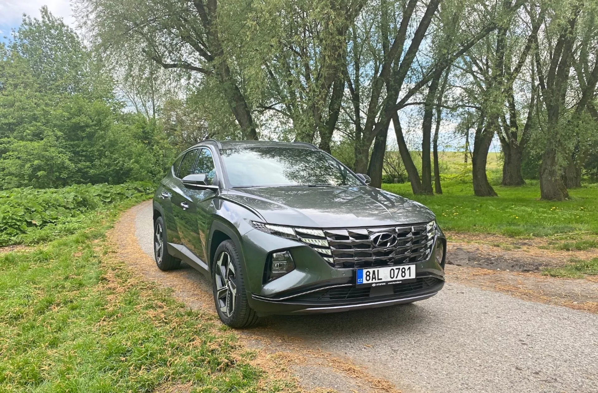 Widok trzy czwarte z przodu przedstawiający Hyundai Tucson 1.6 T-GDi Pure 2020, ukazując nowoczesne cechy SUV jak eleganckie reflektory LED, wyraźnie zaznaczony przedni grill o geometrycznym wzorze, rzeźbiona aerodynamiczna maska, aluminiowe felgi, boczne lusterka z wbudowanymi kierunkowskazami oraz metalizowany lakier, na tle wąskiej asfaltowej drogi otoczonej drzewami, podkreślając jego wszechstronność i atrakcyjność zarówno w warunkach miejskich, jak i wiejskich.