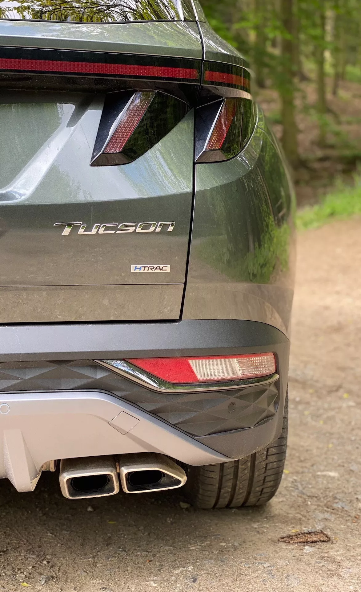 Częściowy widok tylnej części modelu Hyundai Tucson 2020 wskazujący, że jest to wersja 1.6 T-GDi Pure, ukazujący prawą stronę zderzaka, częściową tylną lampę, tylny spojler z LEDowymi światłami hamowania, podwójne końcówki wydechu oraz oznaczenia modelu z nazwą 'Tucson' i technologią silnika 'T-GDi'.