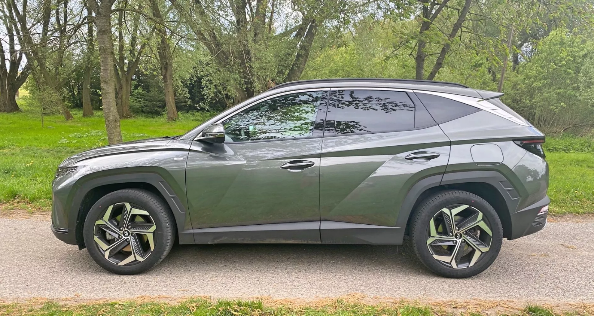 Profil boczny nowoczesnego SUV Hyundai Tucson 1.6 T-GDI Pure 2020 zaparkowanego na utwardzonej drodze obok obszaru trawiastego z drzewami. Prezentuje elegancki, aerodynamiczny design ze schodzącą linią dachu, charakterystyczną linią nadwozia, dużymi, wieloszprychowymi felgami aluminiowymi z oponami o niskim profilu, przyciemnianymi szybami z widocznym słupkiem B, wpuszczonymi w nadwozie klamkami, kolorowymi lusterkami bocznymi i chromowaną listwą wzdłuż krawędzi okna. Pojazd ukazuje współczesny styl i możliwe zaawansowane funkcje z kąta bocznego.